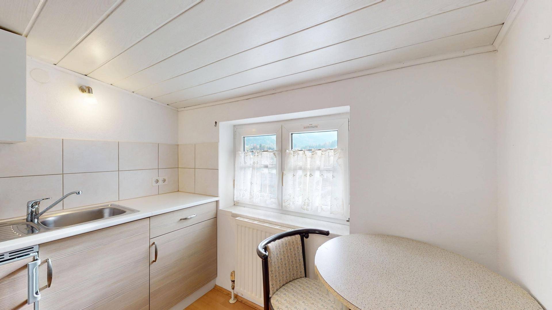 Kleine Küchenzeile mit Spüle, Unterschränken und Fenster mit Vorhängen.