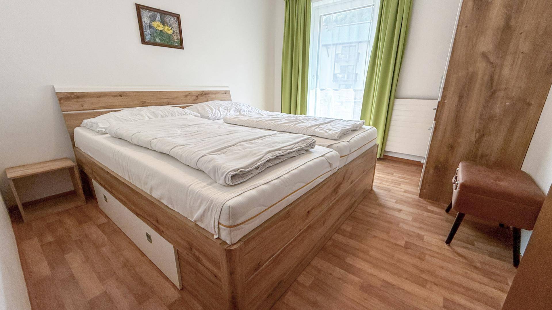 Schlafzimmer mit Doppelbett aus Holz, Nachttisch und grünen Vorhängen am Fenster.