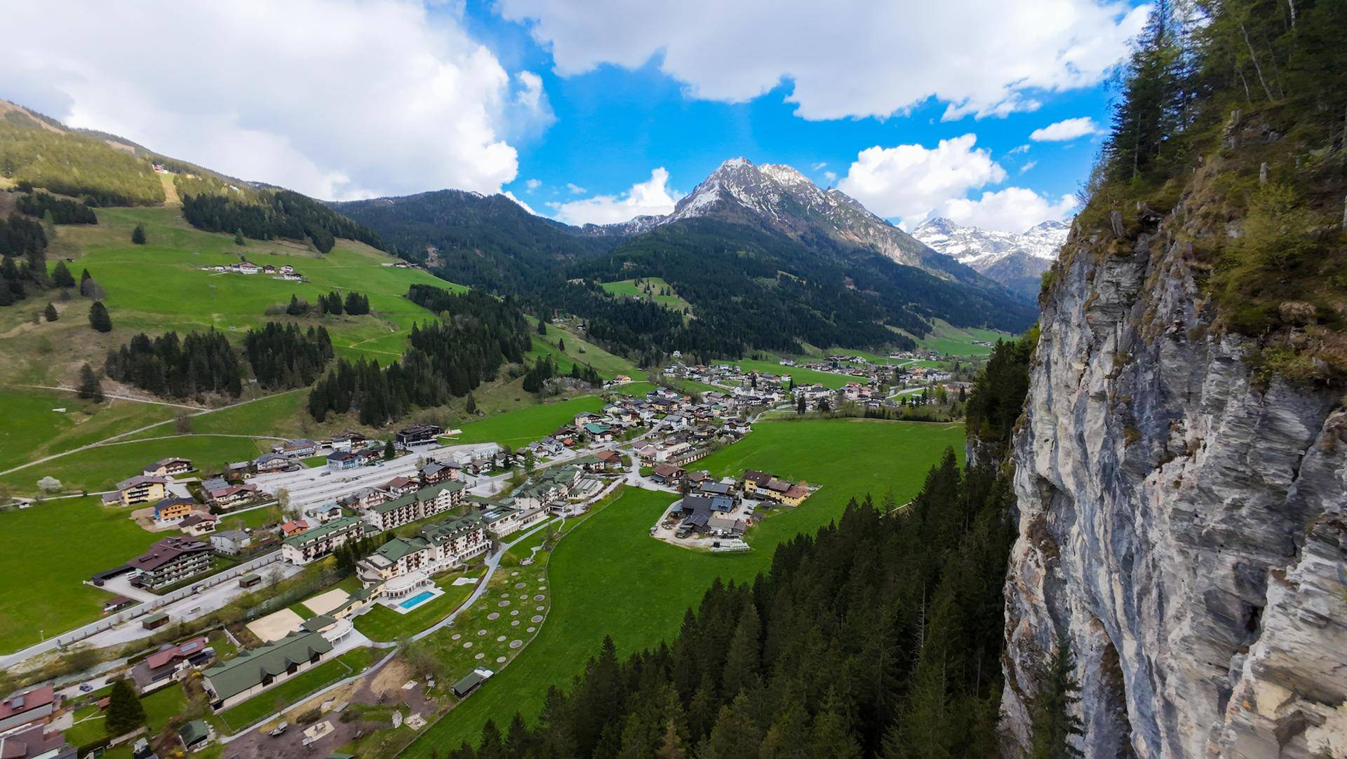Weitläufige Ansicht eines Bergdorfes in einem Tal mit grünen Wiesen und felsigen Klippen.