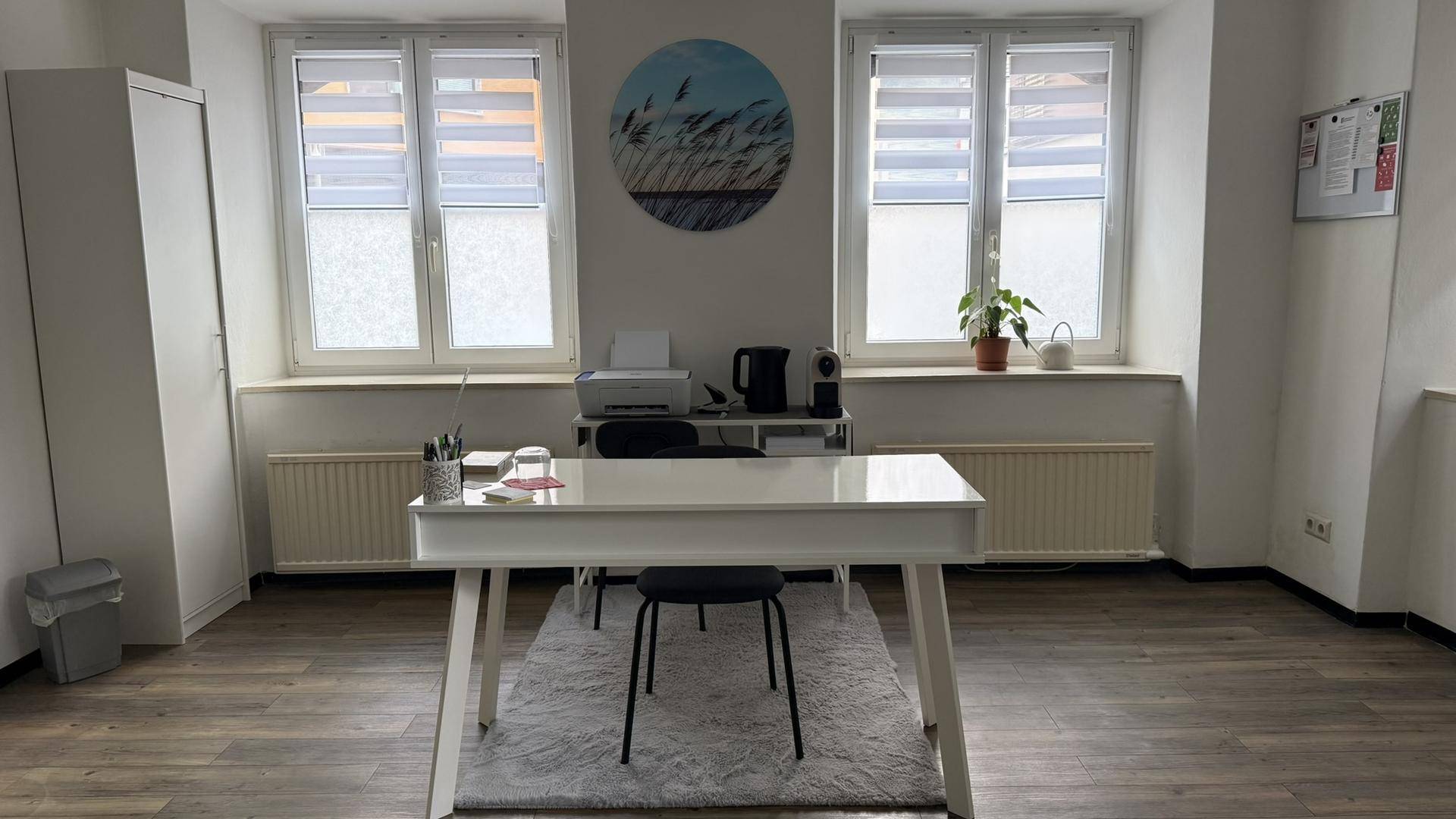 Detailansicht des Schreibtischbereichs mit Drucker, Kaffeemaschine und zwei großen Fenstern.