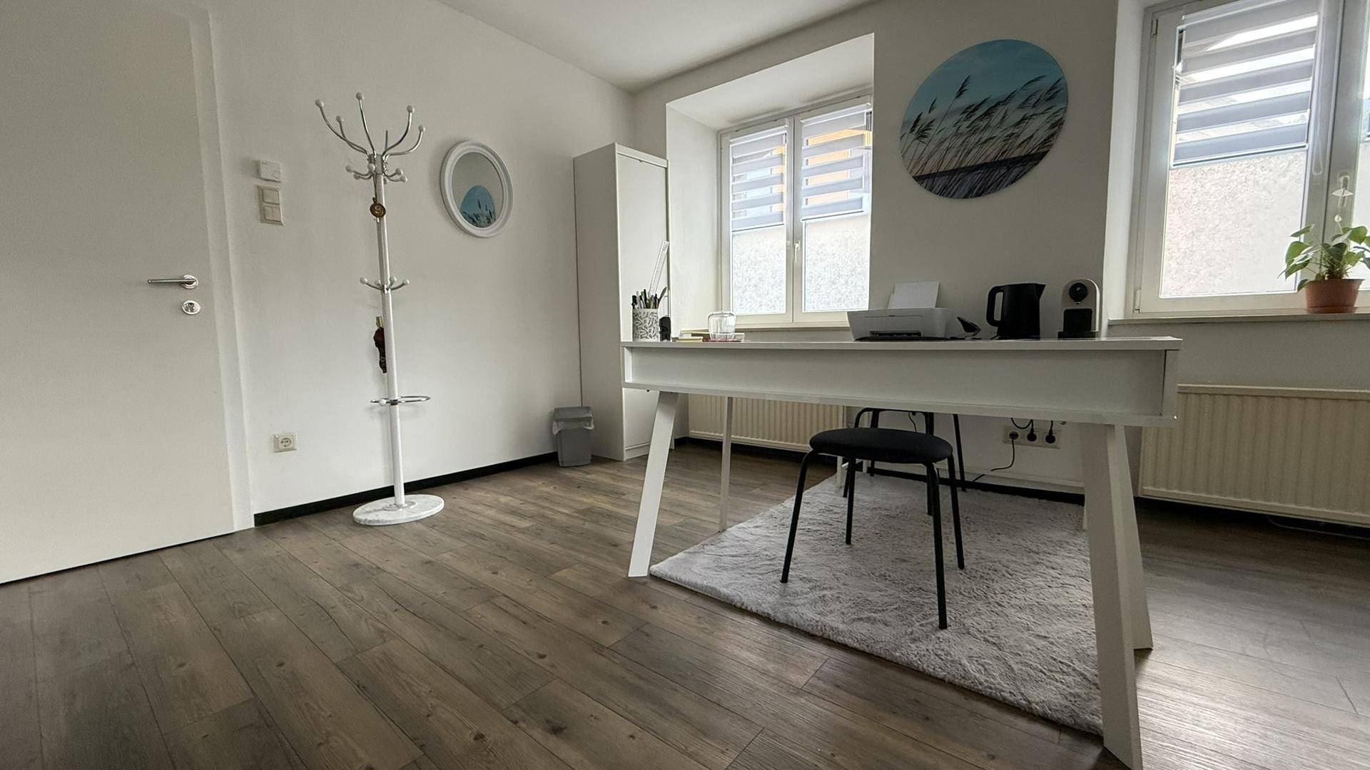 Büroraum mit Schreibtisch, Garderobe und Fenstern, die für eine angenehme Arbeitsatmosphäre sorgen.