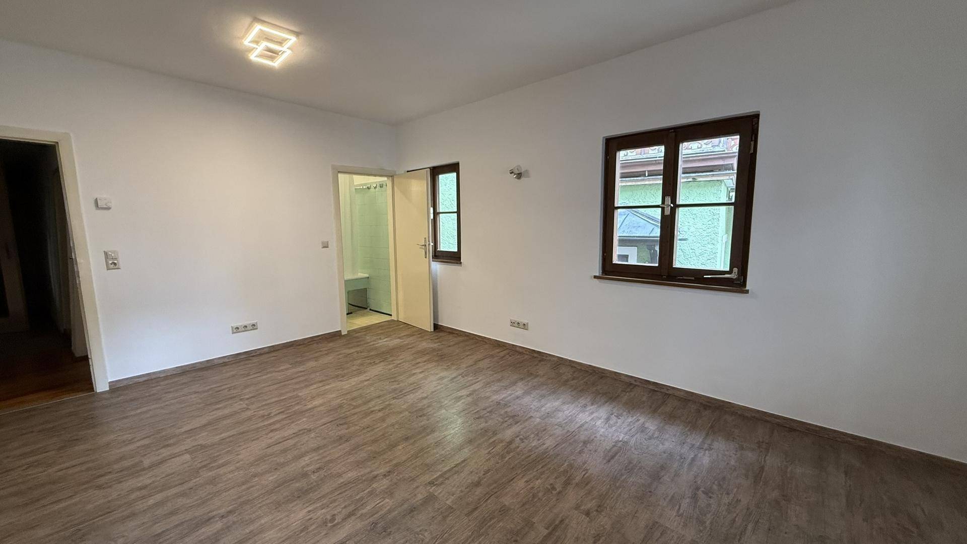 Geräumiger Wohnbereich mit Laminatboden, Fenstern und Zugang zu einem weiteren Zimmer.