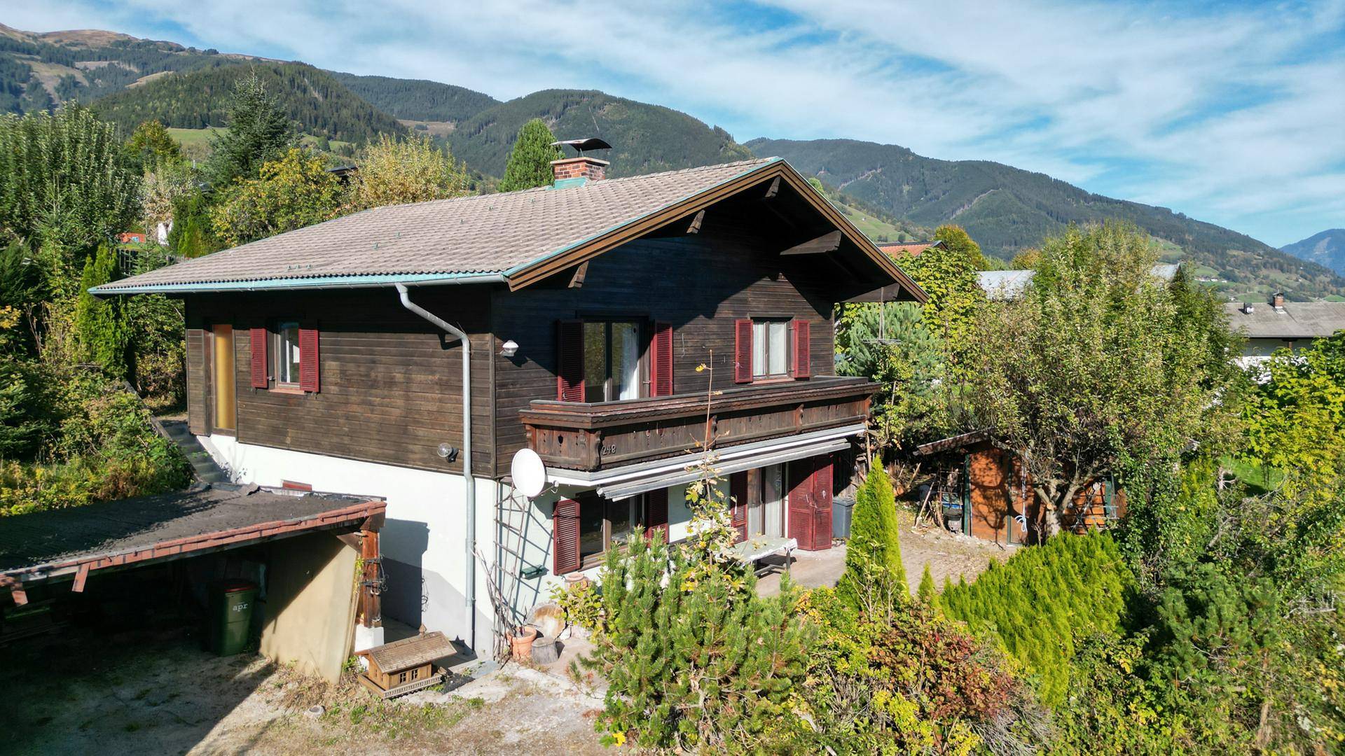Freistehendes Holzhaus mit Satteldach, Balkon und Carport, eingebettet in eine grüne Hügellandschaft.