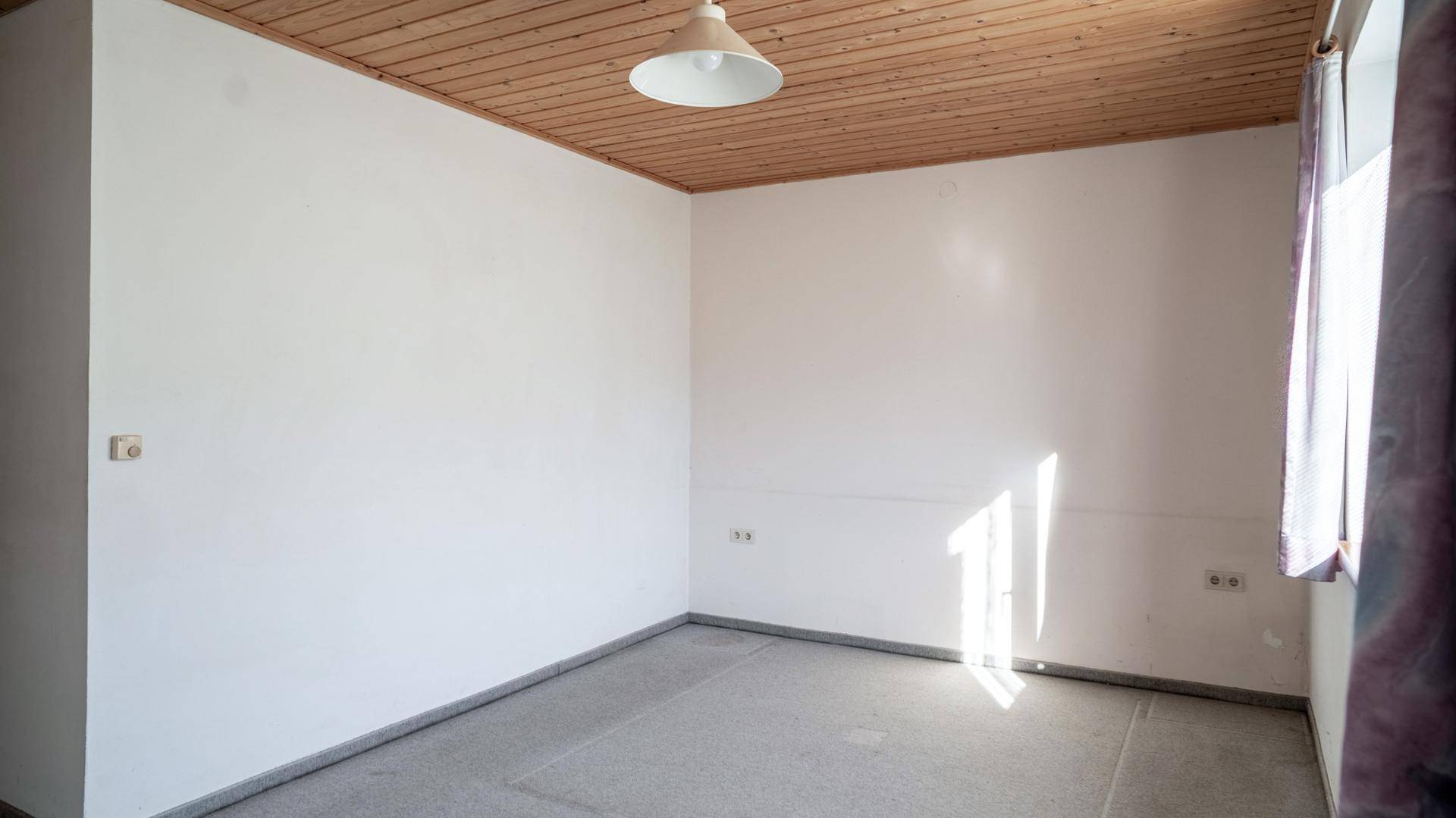 Heller, leerer Raum mit weißen Wänden, einer Holzdecke und grauem Bodenbelag. Natürliches Licht durchflutet den Raum.