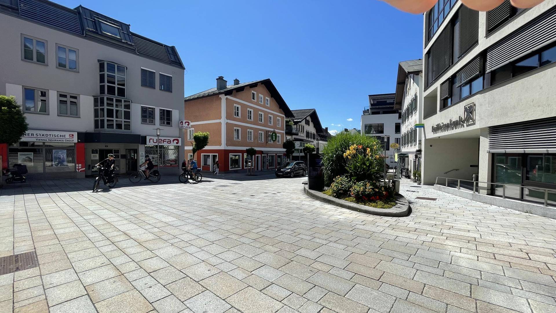 Belebter Stadtplatz mit verschiedenen Gebäuden, gepflastertem Boden und sonnigem Wetter.