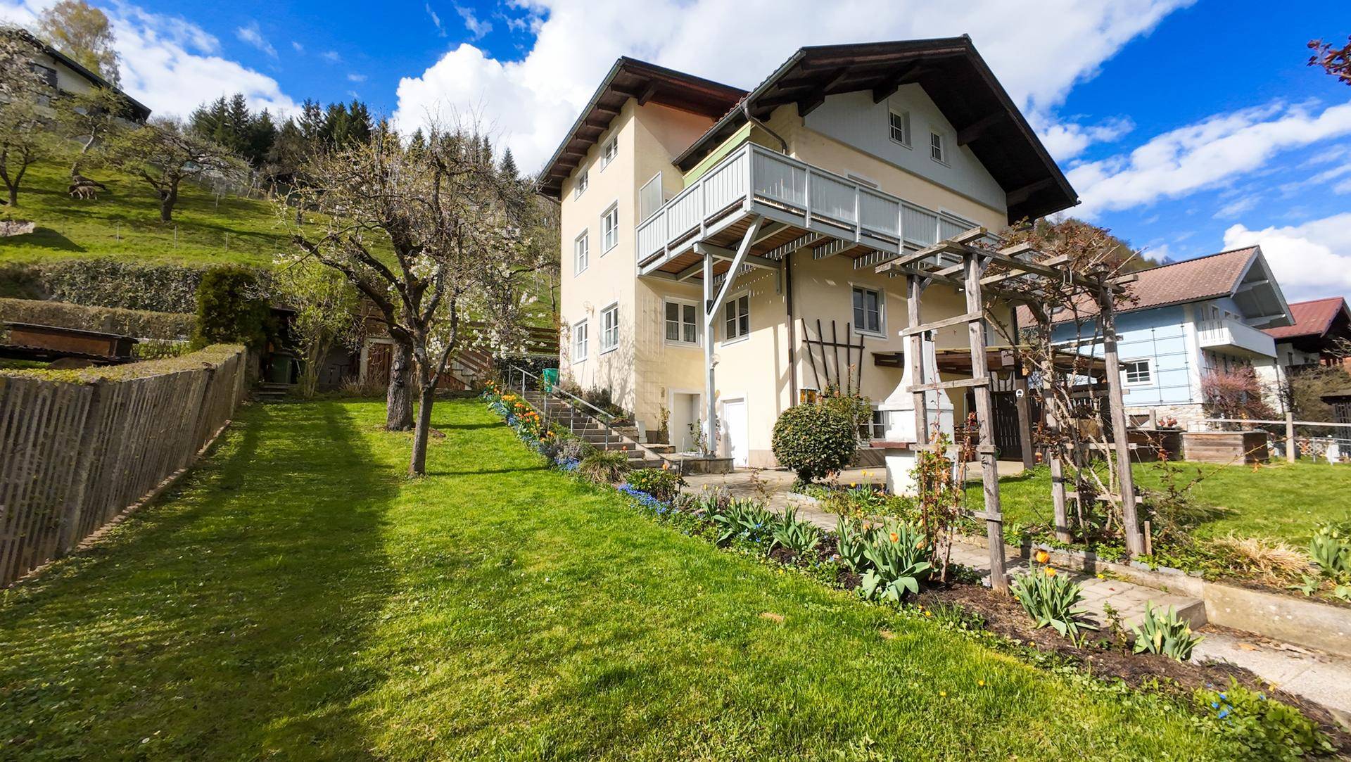 Weitläufiger Gartenbereich mit Rasen, Blumenbeeten und Blick auf das Haus und die Berge.