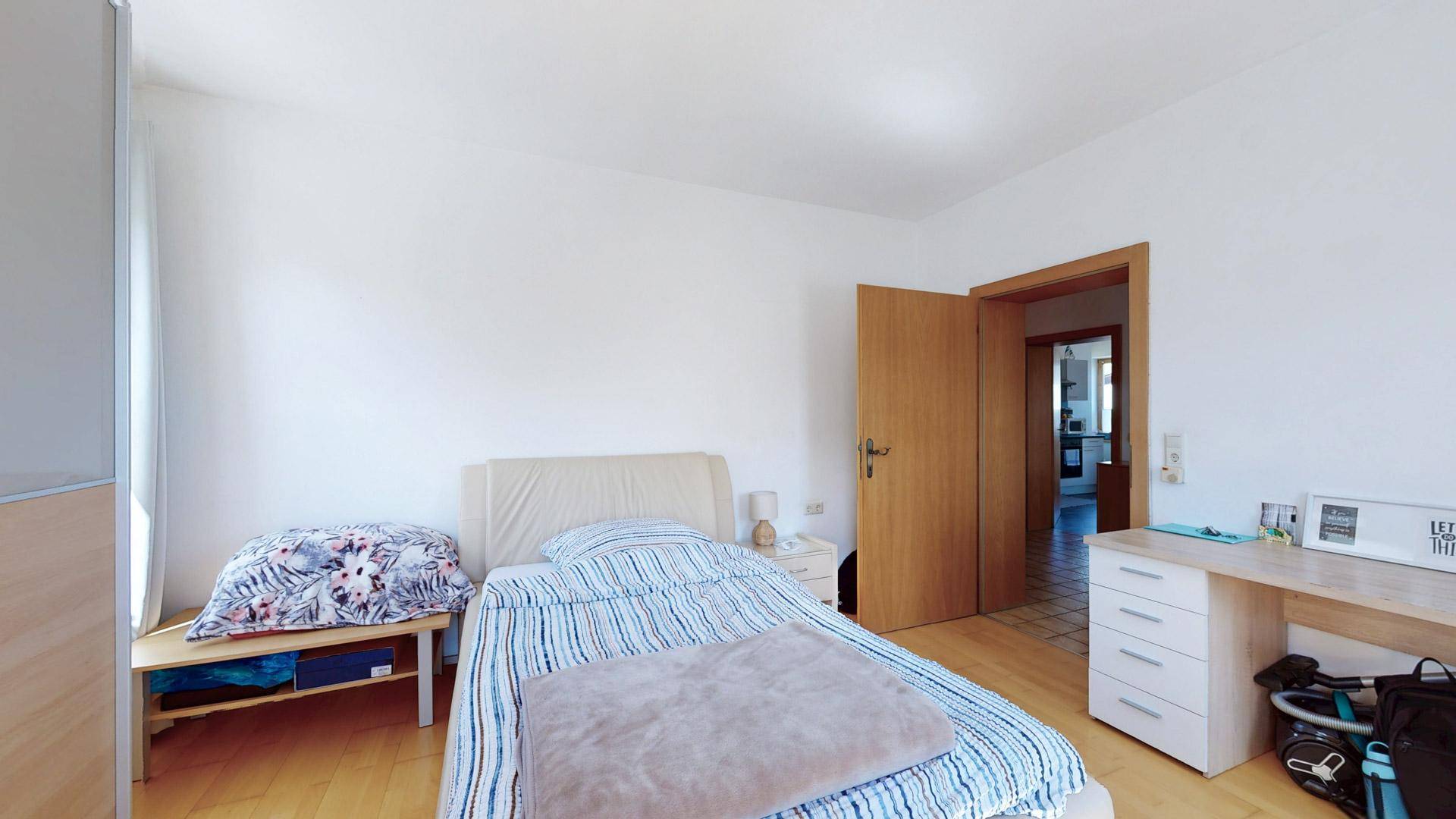 Helles Schlafzimmer mit Bett, Schreibtisch und Zugang zu einem weiteren Raum.