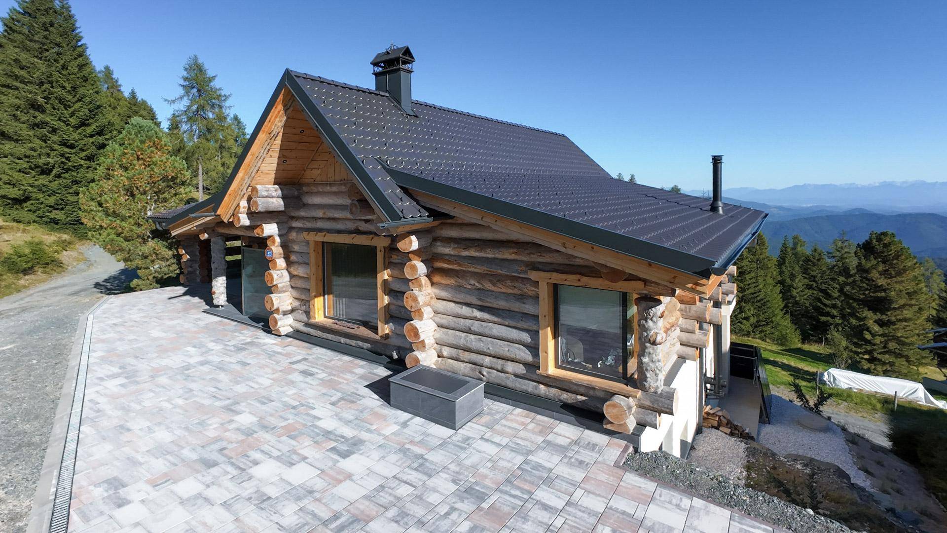 Blockhaus mit Holzbalken und gepflastertem Vorplatz, umgeben von Nadelbäumen und Berglandschaft.