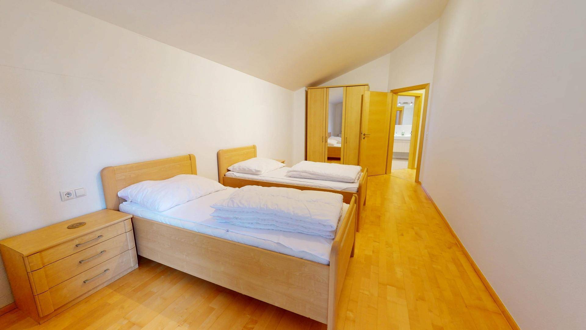Helles Schlafzimmer mit zwei Einzelbetten, Holzboden und Zugang zum Badezimmer.