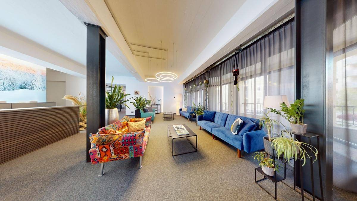 Einladender Loungebereich mit farbenfrohen Sofas, vielen Pflanzen und großen Fenstern, die viel Tageslicht hereinlassen.