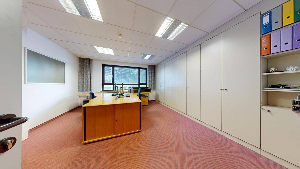 Büro mit Teppichboden, großen Fenstern und integrierten Schränken, die viel Stauraum bieten.
