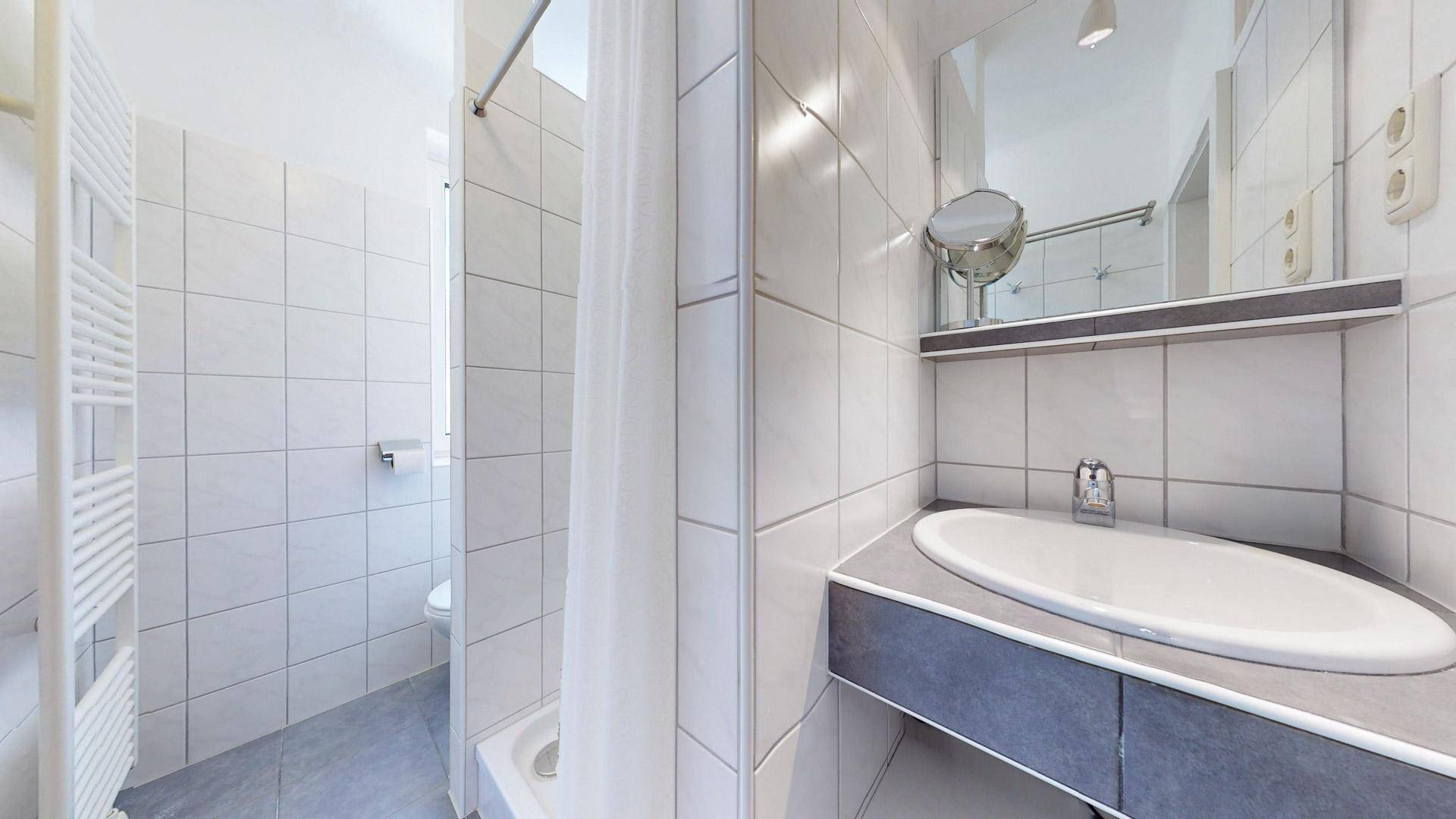 Modernes Badezimmer mit Duschbereich, Waschtisch und hellen Fliesen an Wänden und Boden.