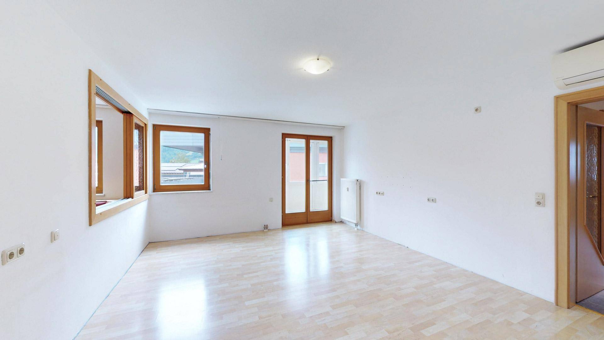 Leeres Wohnzimmer mit hellem Holzboden, weißen Wänden und Zugang zu einem Außenbereich.