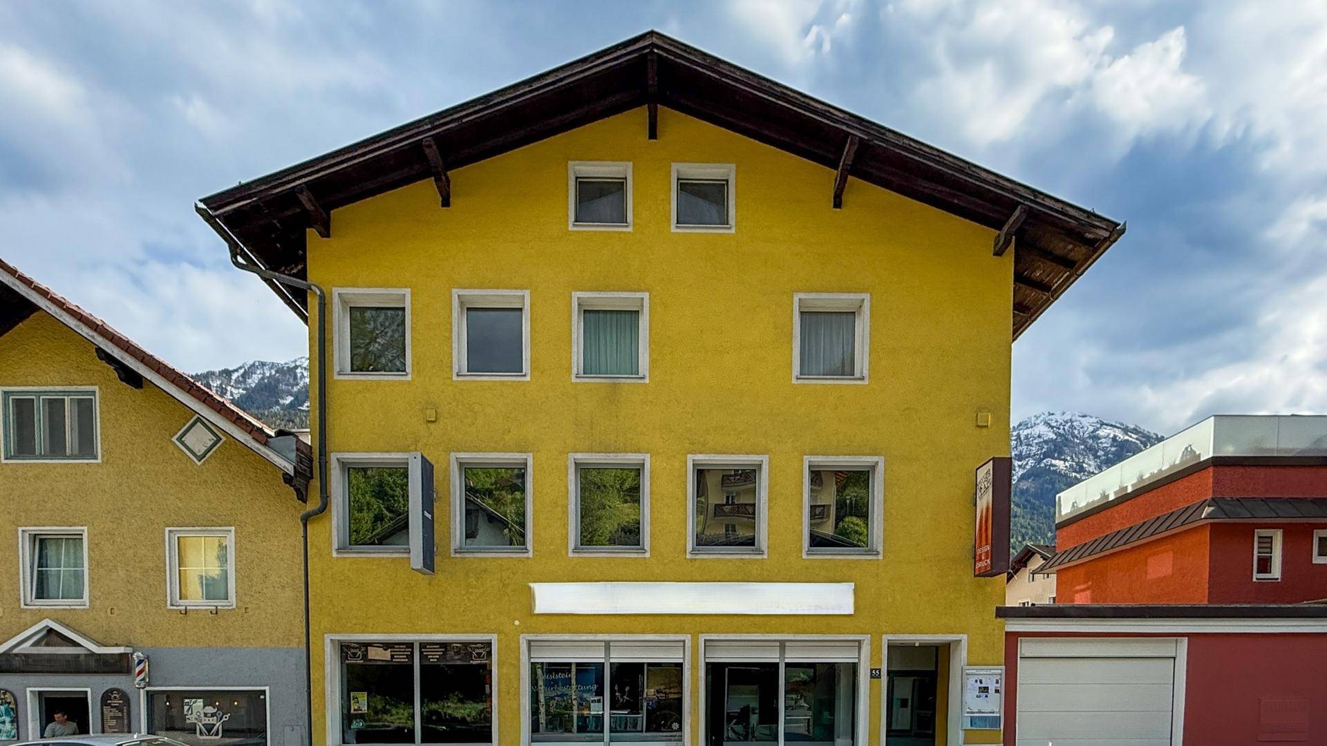 Gelbes Gebäude mit traditioneller Architektur und großen Fenstern, umgeben von Bergen und blauem Himmel.
