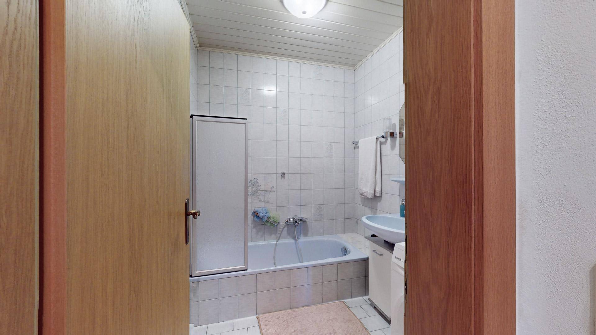 Badezimmer mit Badewanne, Duschabtrennung und Waschbecken, komplett gefliest in Weiß.