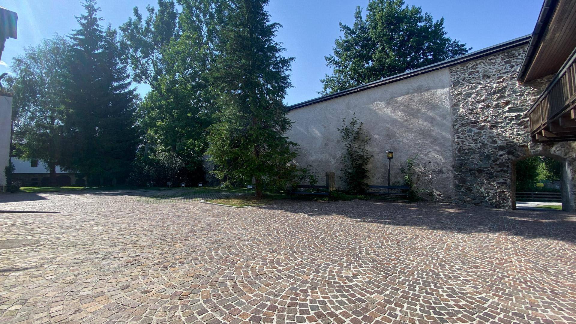 Großer gepflasterter Innenhof mit Kopfsteinpflaster, umgeben von alten Steinmauern und Bäumen.