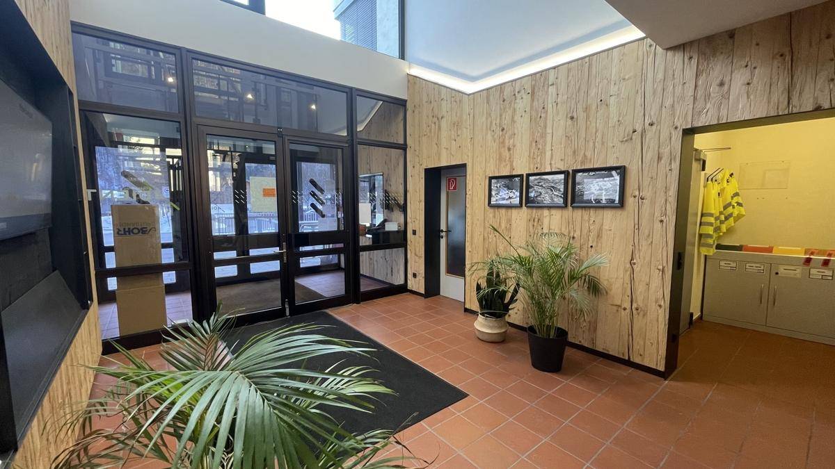 Heller Eingangsbereich mit großen Glasfronten, Holzverkleidung und Fliesenboden, der zu weiteren Räumen führt.