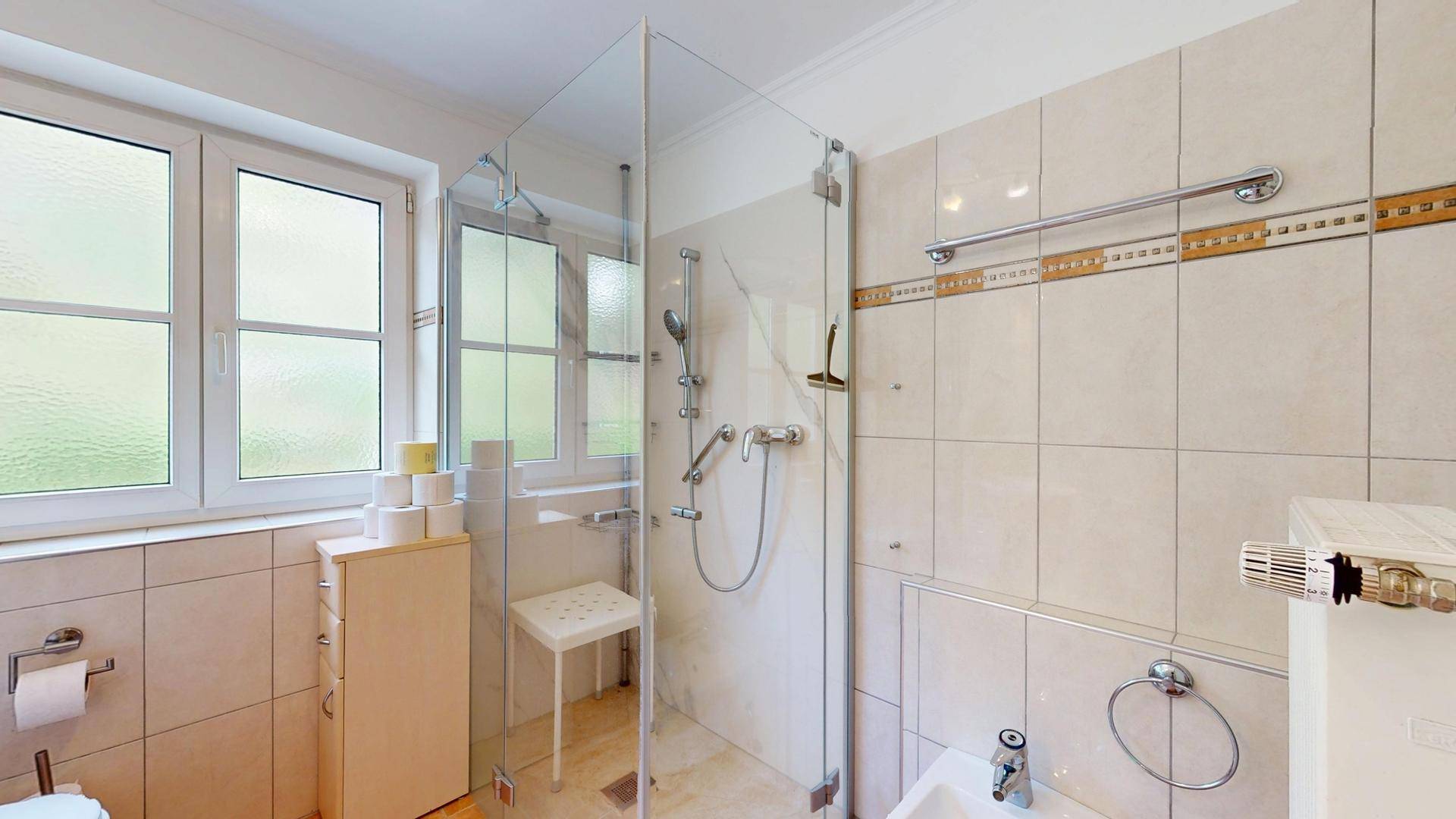 Modernisiertes Badezimmer mit ebenerdiger Dusche, Fenster und hellen Fliesen.