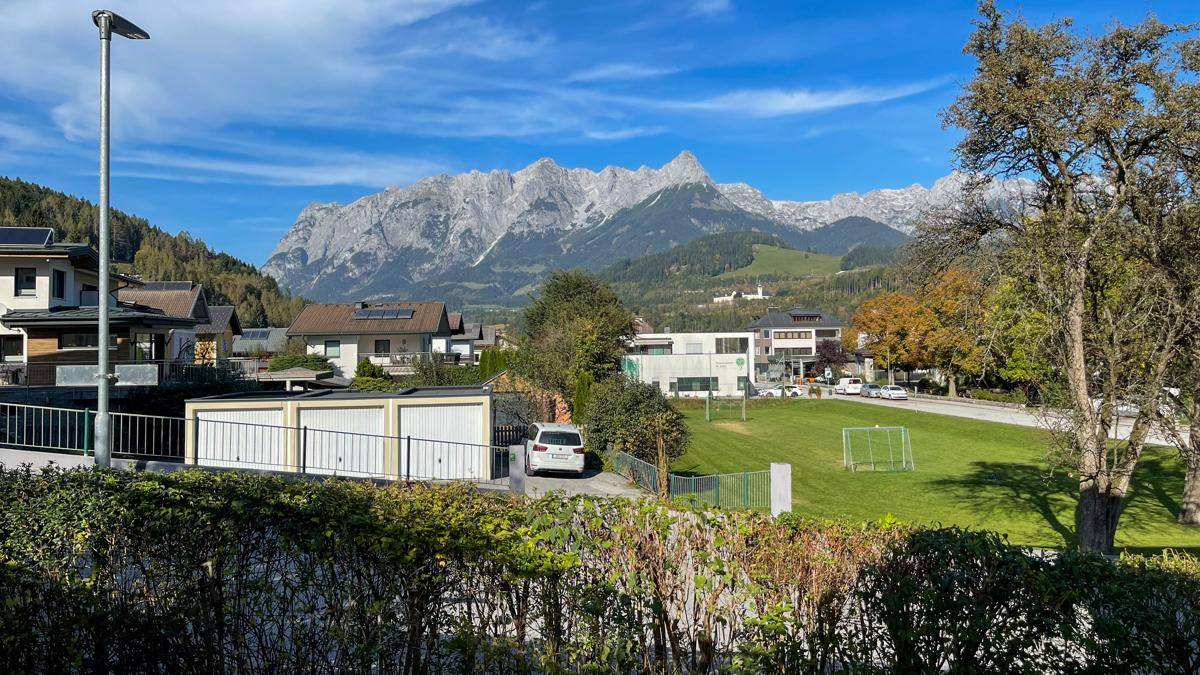 Malerische Aussicht auf eine Stadt mit grünen Wiesen, Häusern und imposanten Bergen im Hintergrund unter blauem Himmel.