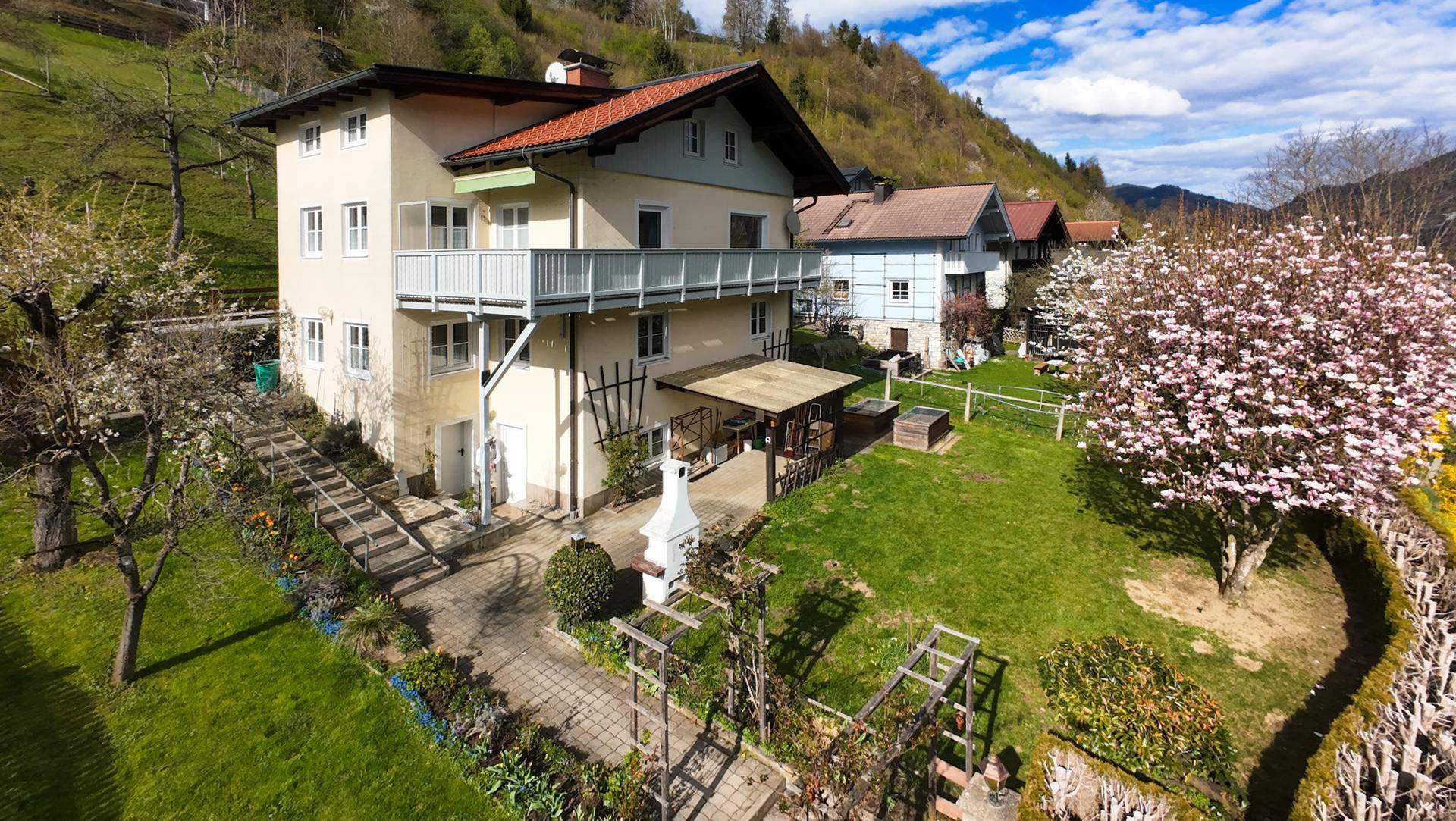 Panoramablick auf das Haus und den weitläufigen Garten mit blühenden Bäumen und Bergblick.