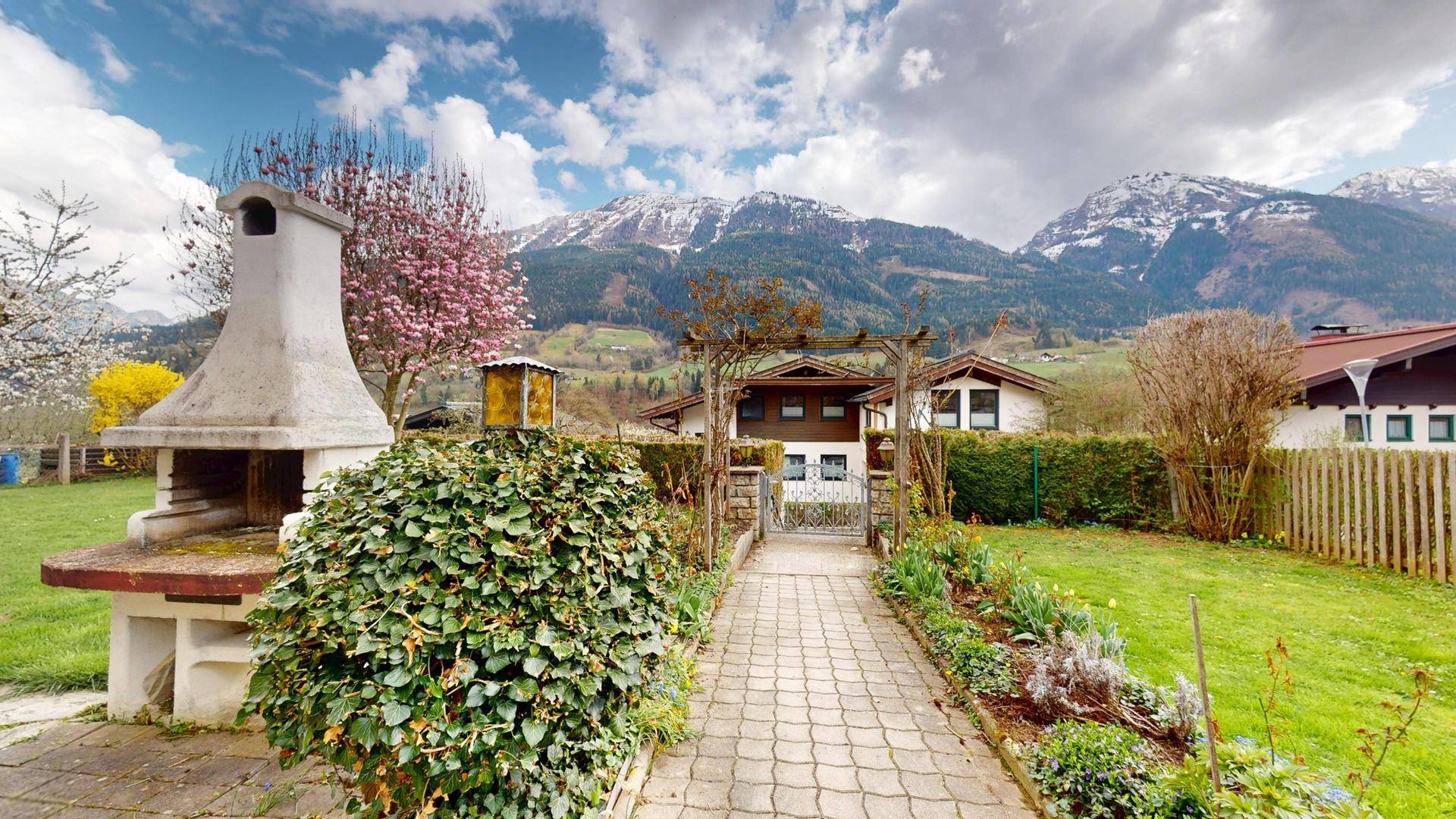 Gartenweg führt durch blühende Beete zu einem Tor mit Bergblick im Hintergrund.
