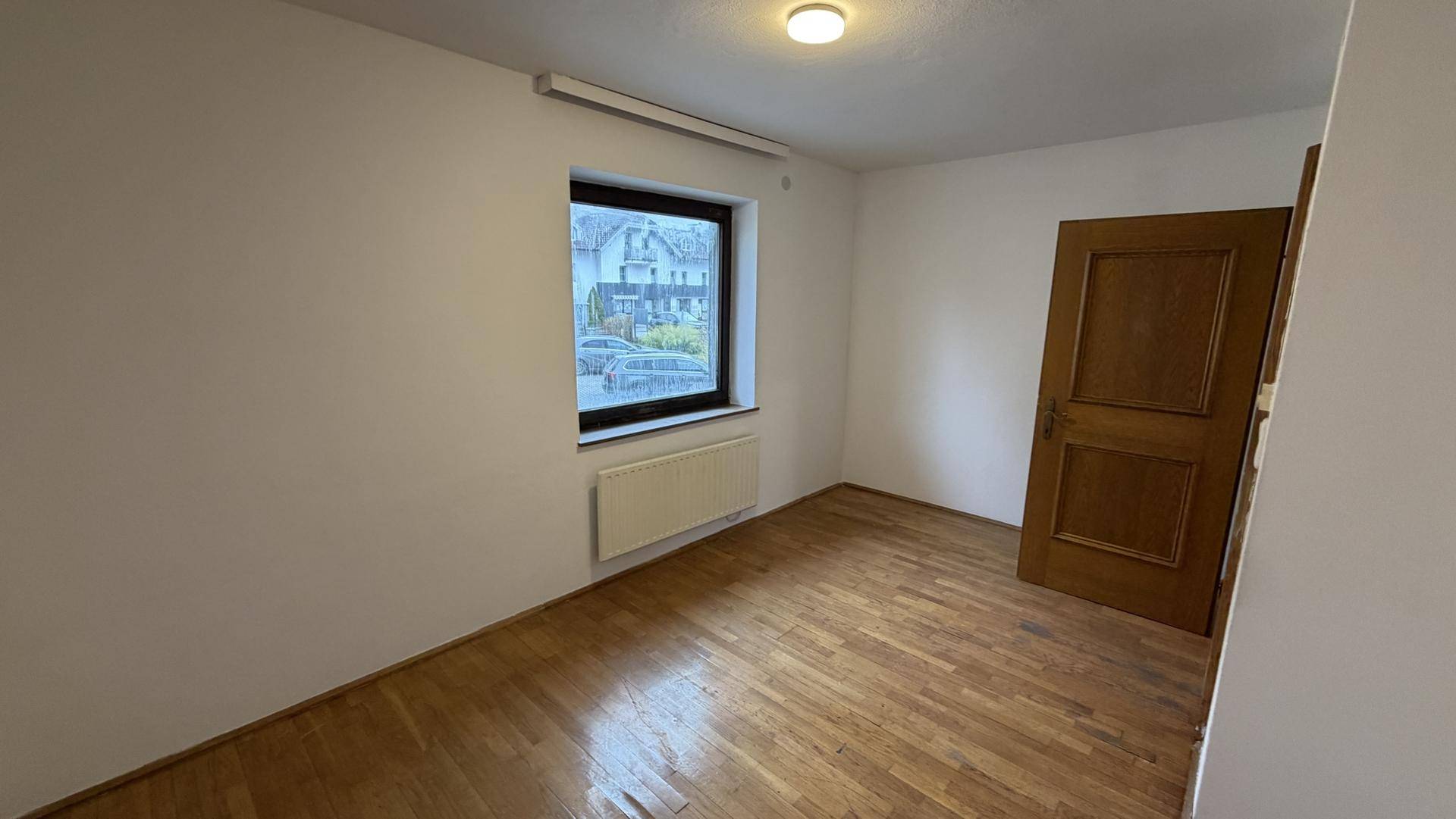 Helles Zimmer mit Holzparkett, Heizkörper und Fenster mit Blick nach draußen.