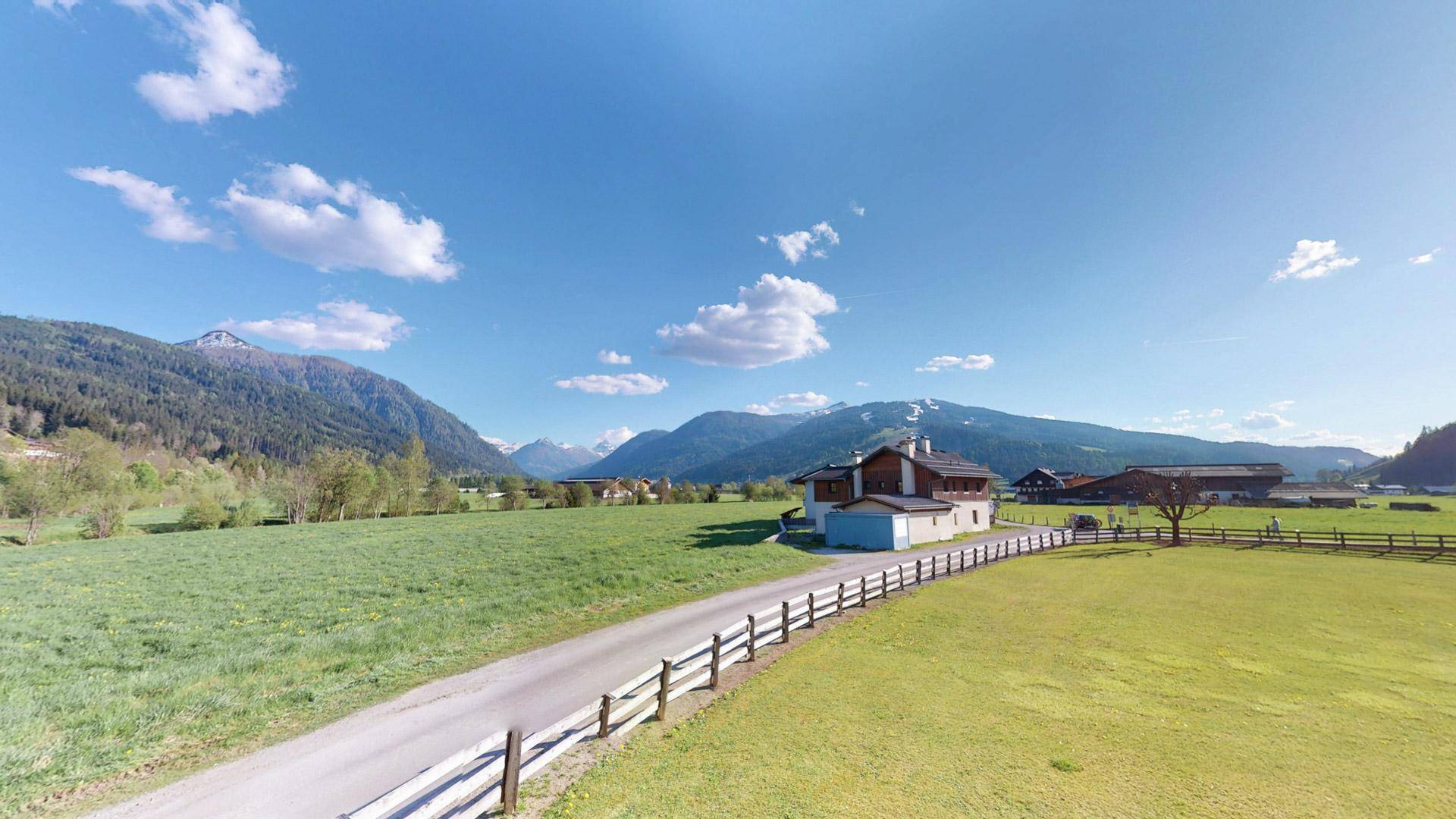 Weitläufige Landschaft mit einem Haus im Grünen und Bergen im Hintergrund unter blauem Himmel.