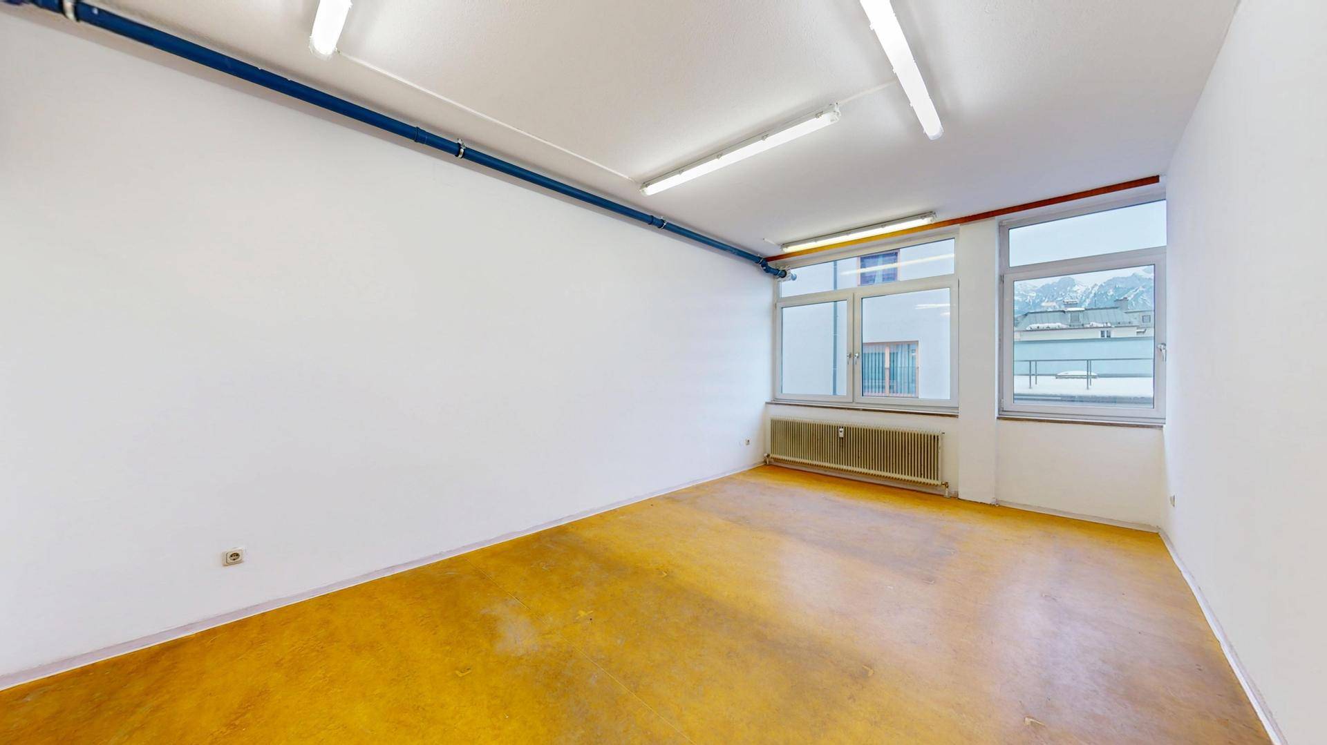 Leerer Büroraum mit gelbem Boden, weißen Wänden und sichtbaren Rohren an der Decke.