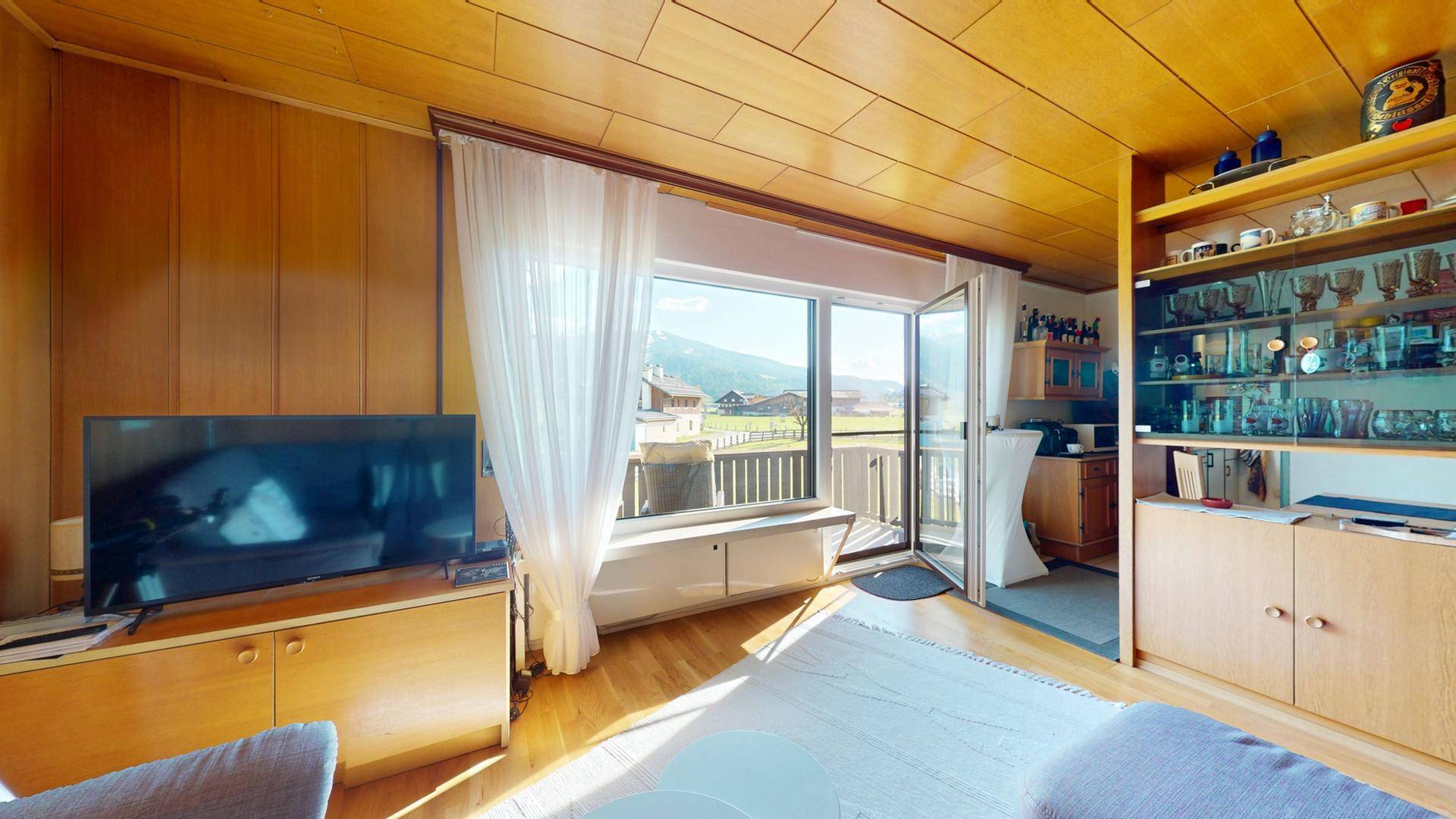 Gemütliches Wohnzimmer mit Holzboden, TV-Möbel und Zugang zum Balkon mit Blick ins Grüne.
