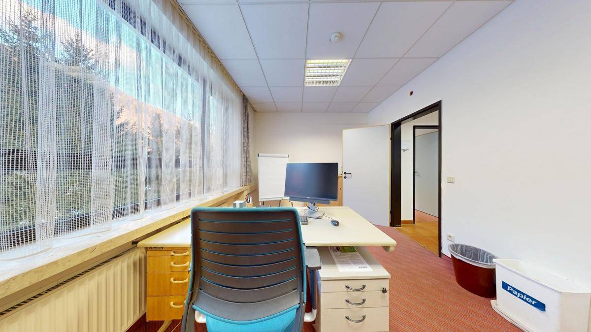 Büro mit Teppichboden, großem Fenster und Schreibtisch, ausgestattet für effizientes Arbeiten.