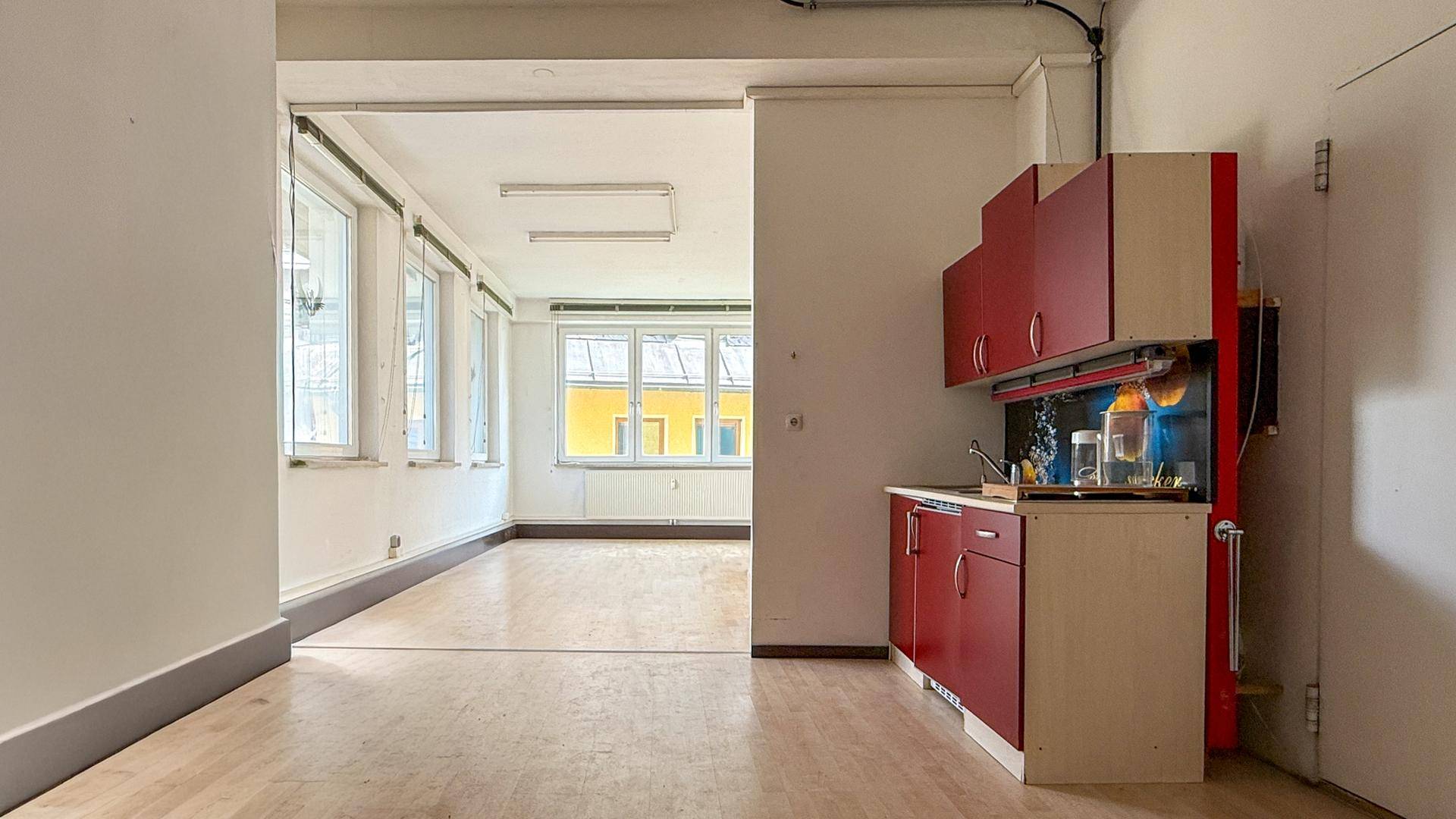 Leerer Büroraum mit Holzboden und einer kleinen Küchenzeile mit roten Schränken, bereit zur Nutzung.