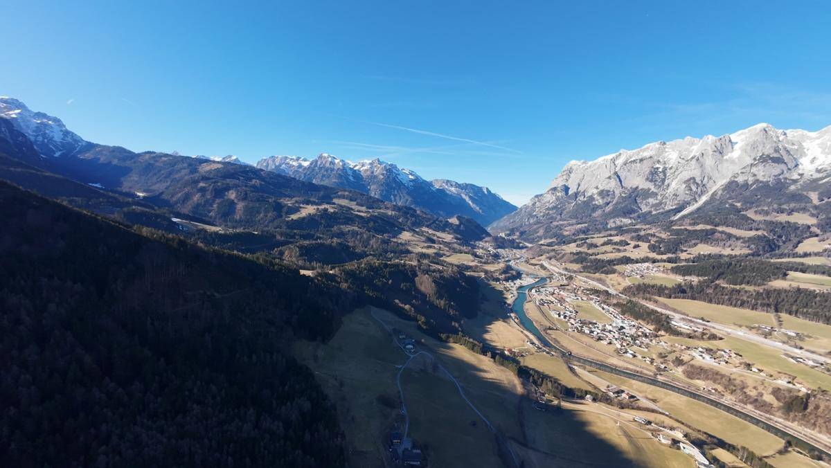 Luftaufnahme eines Flusstals mit schneebedeckten Bergen, einer Siedlung und einer Straße unter klarem Himmel.