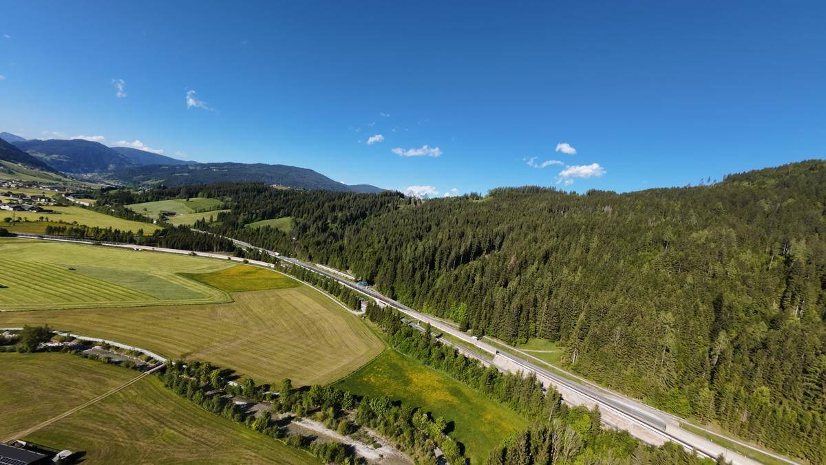 Luftaufnahme einer Autobahn, die sich durch eine bewaldete Berglandschaft mit weiten Feldern schlängelt.