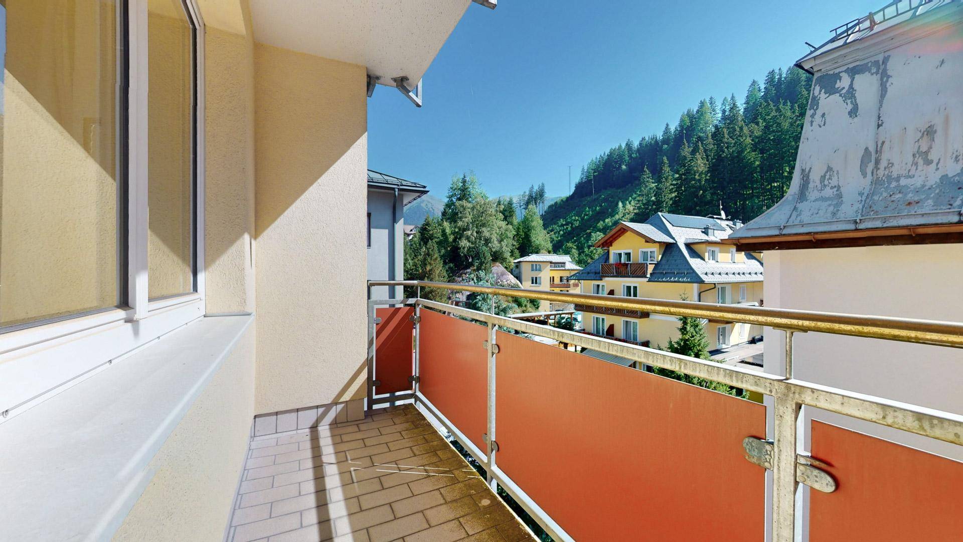 Sonniger Balkon mit orangefarbener Brüstung und Blick auf die umliegenden Häuser und Berge.