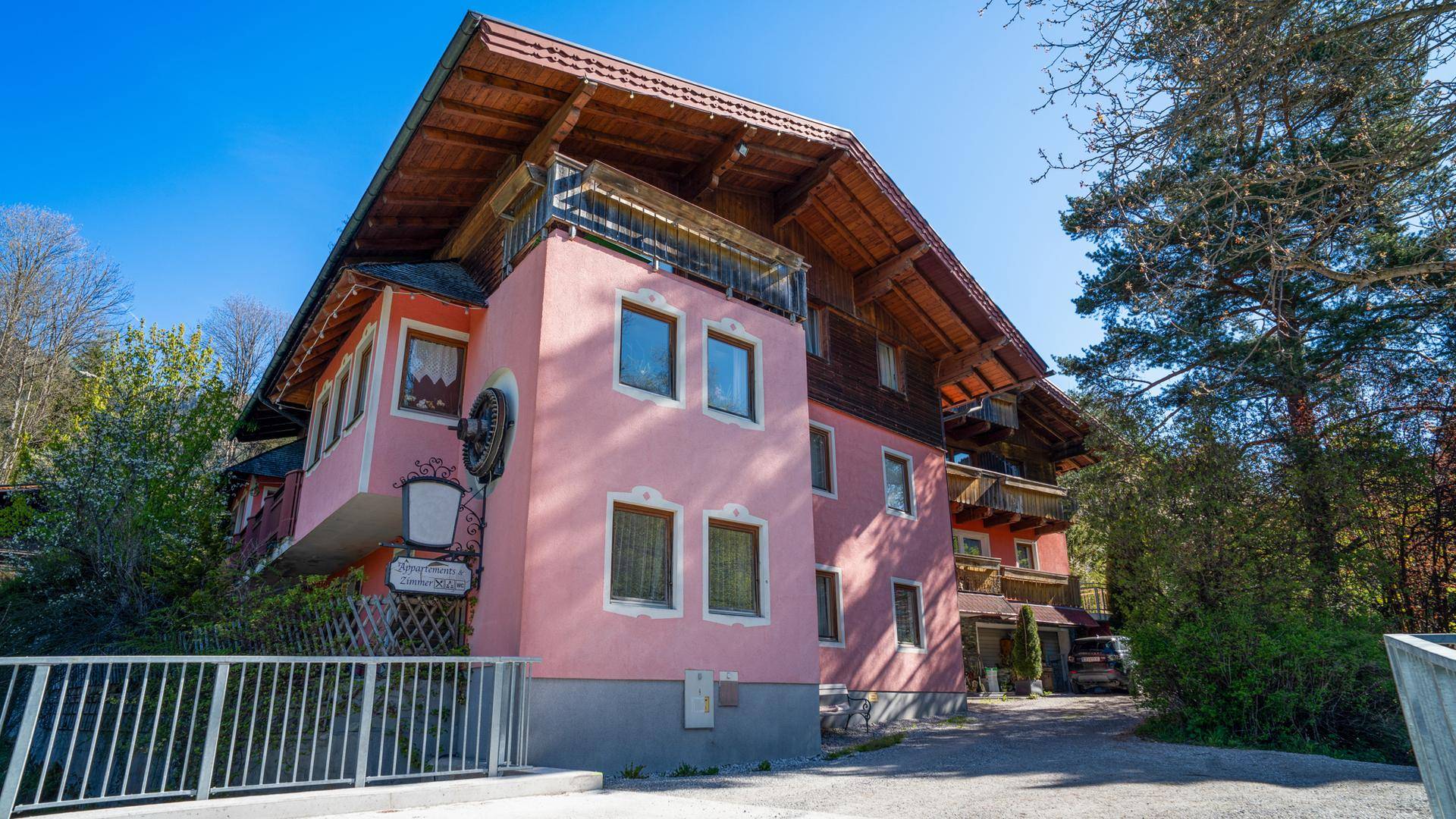 Rosa gestrichenes Gebäude mit Holzbalkonen und traditionellem Dach, umgeben von üppiger Vegetation.
