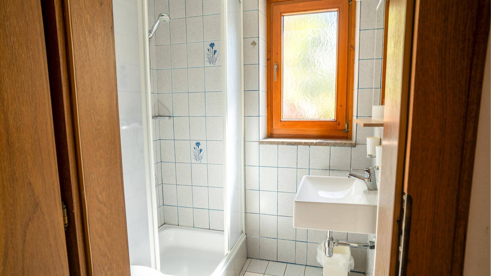 Helles Badezimmer mit Duschkabine, Waschbecken und Fenster, weiß gefliest.