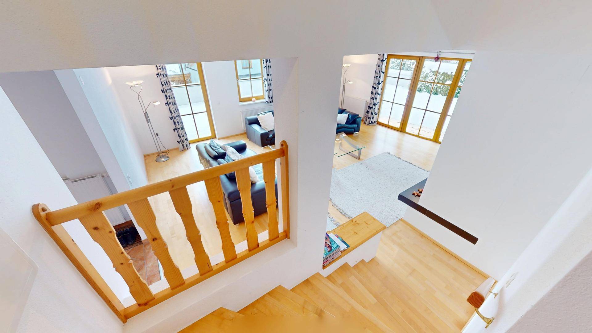 Blick von der Holztreppe hinunter in den hellen Wohnbereich mit großen Fenstern.