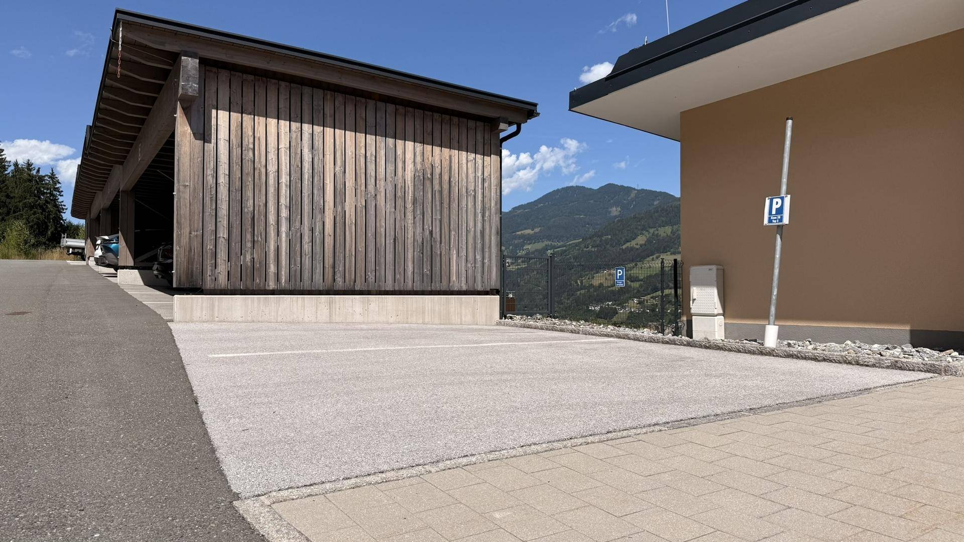 Parkplatzbereich vor einem modernen Gebäude mit Holzverkleidung und Bergblick unter strahlendem Himmel.