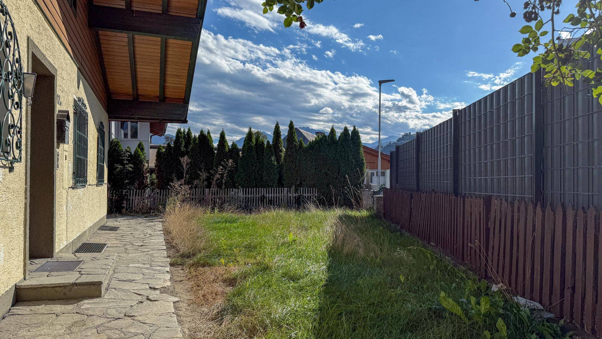 Gartenbereich neben dem Haus mit Rasenfläche, Steinplattenweg und Blick auf die bewaldeten Berge im Hintergrund.