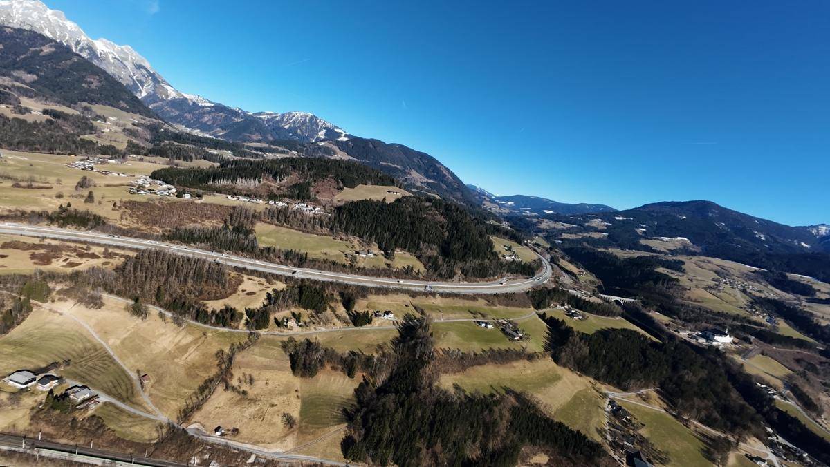 Luftaufnahme eines bewaldeten Tals mit einer kurvigen Autobahn und schneebedeckten Bergen im Hintergrund.