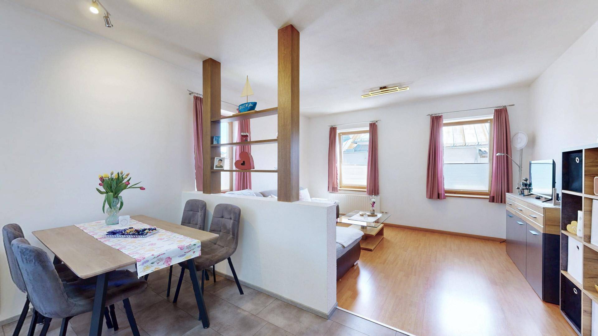 Essbereich mit Holztisch und Stühlen, offen zum Wohnzimmer mit Sofa und Fenstern.