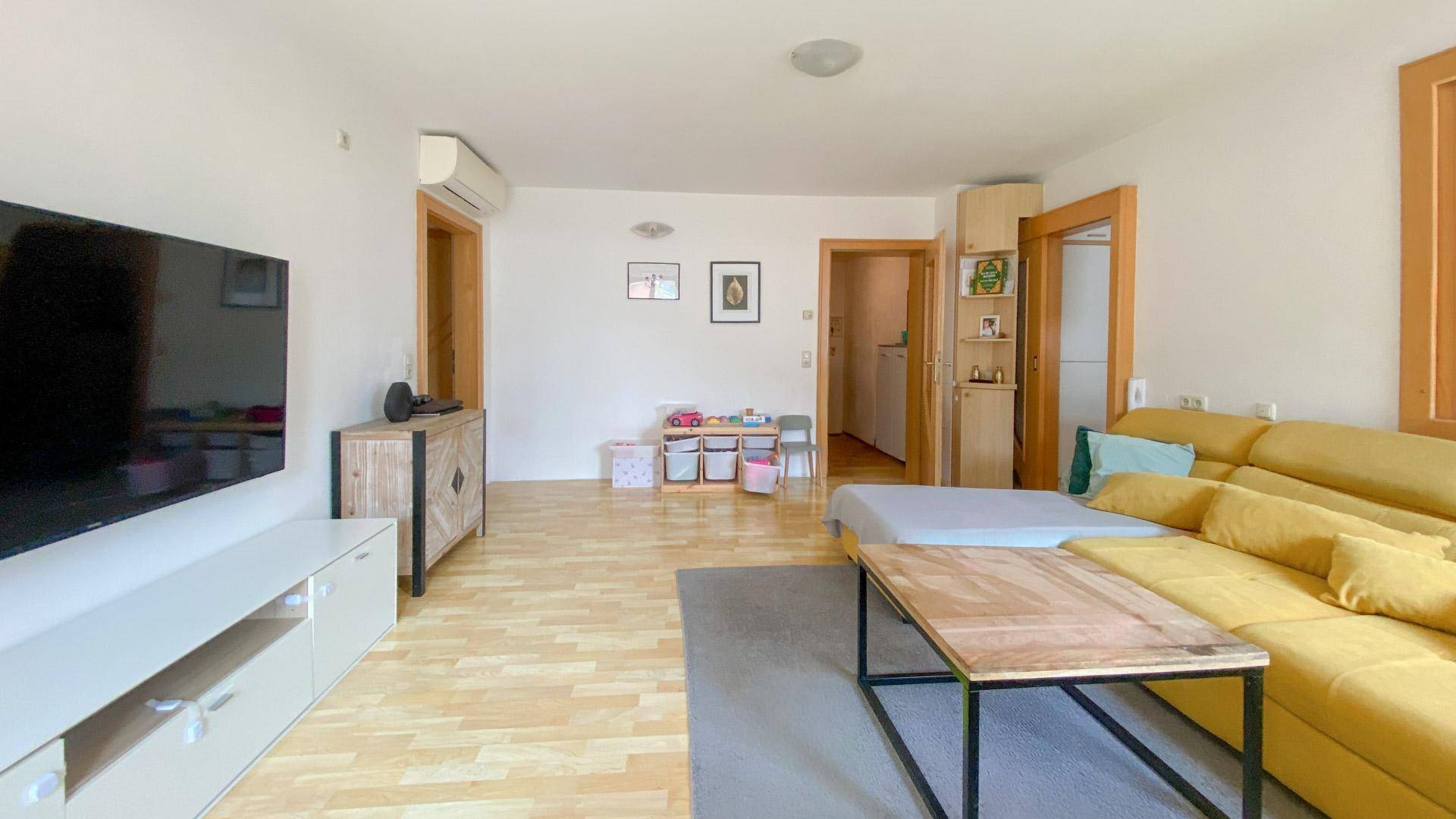 Geräumiges Wohnzimmer mit Holzboden, Klimaanlage und Blick auf den Eingangsbereich.