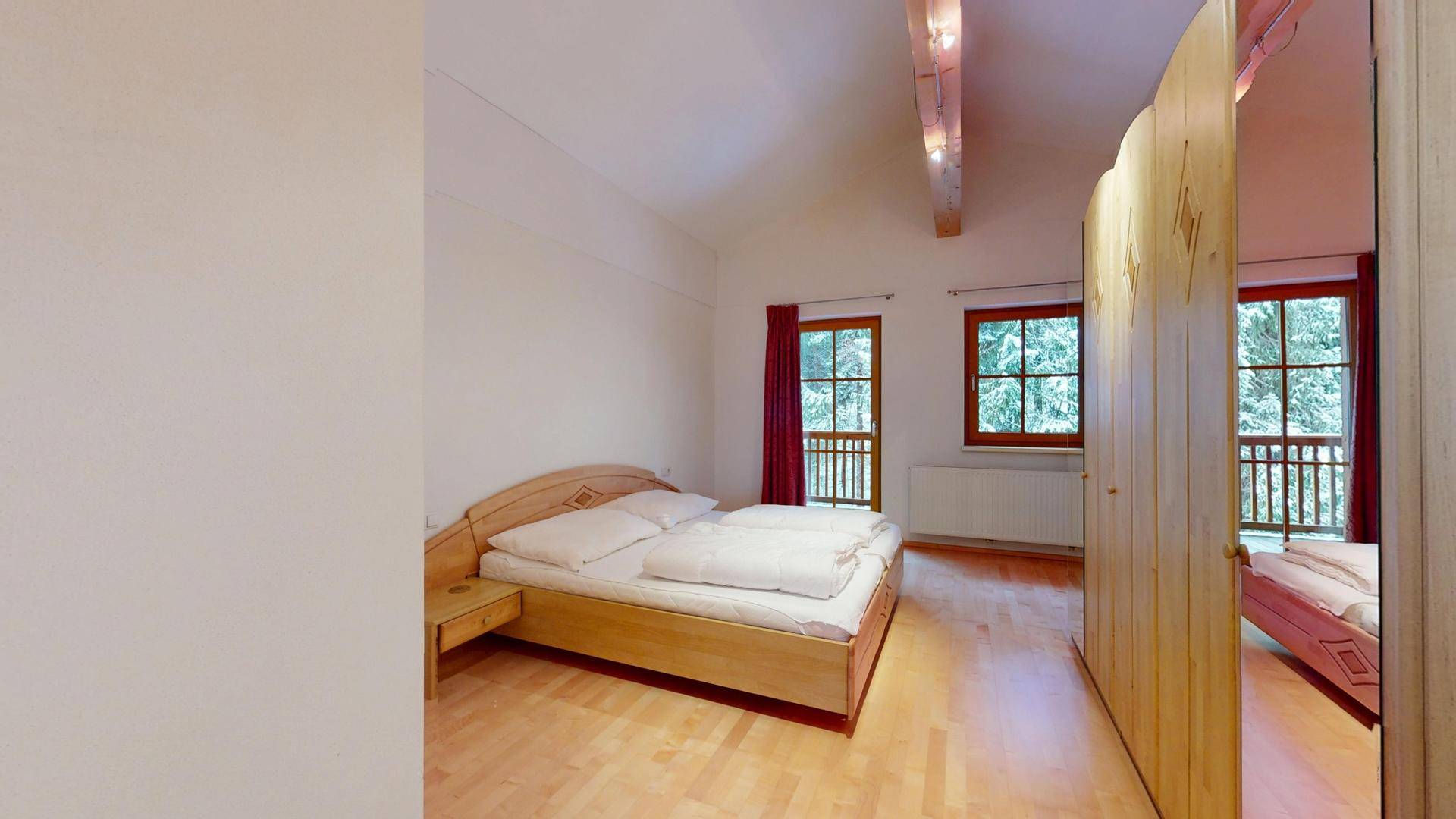 Schlafzimmer mit Holzbett, Parkettboden und Fenstern mit Blick auf verschneite Bäume.