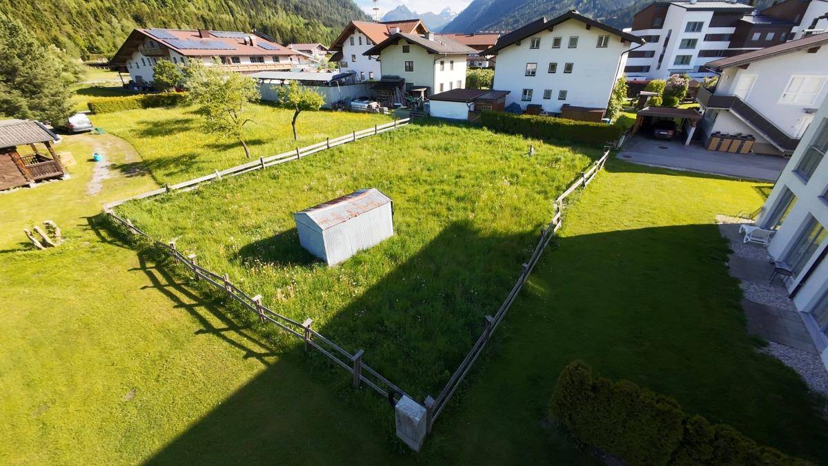 Grünes Grundstück mit einem Schuppen, umzäunt und umgeben von Wohnhäusern und Bergen im Hintergrund.
