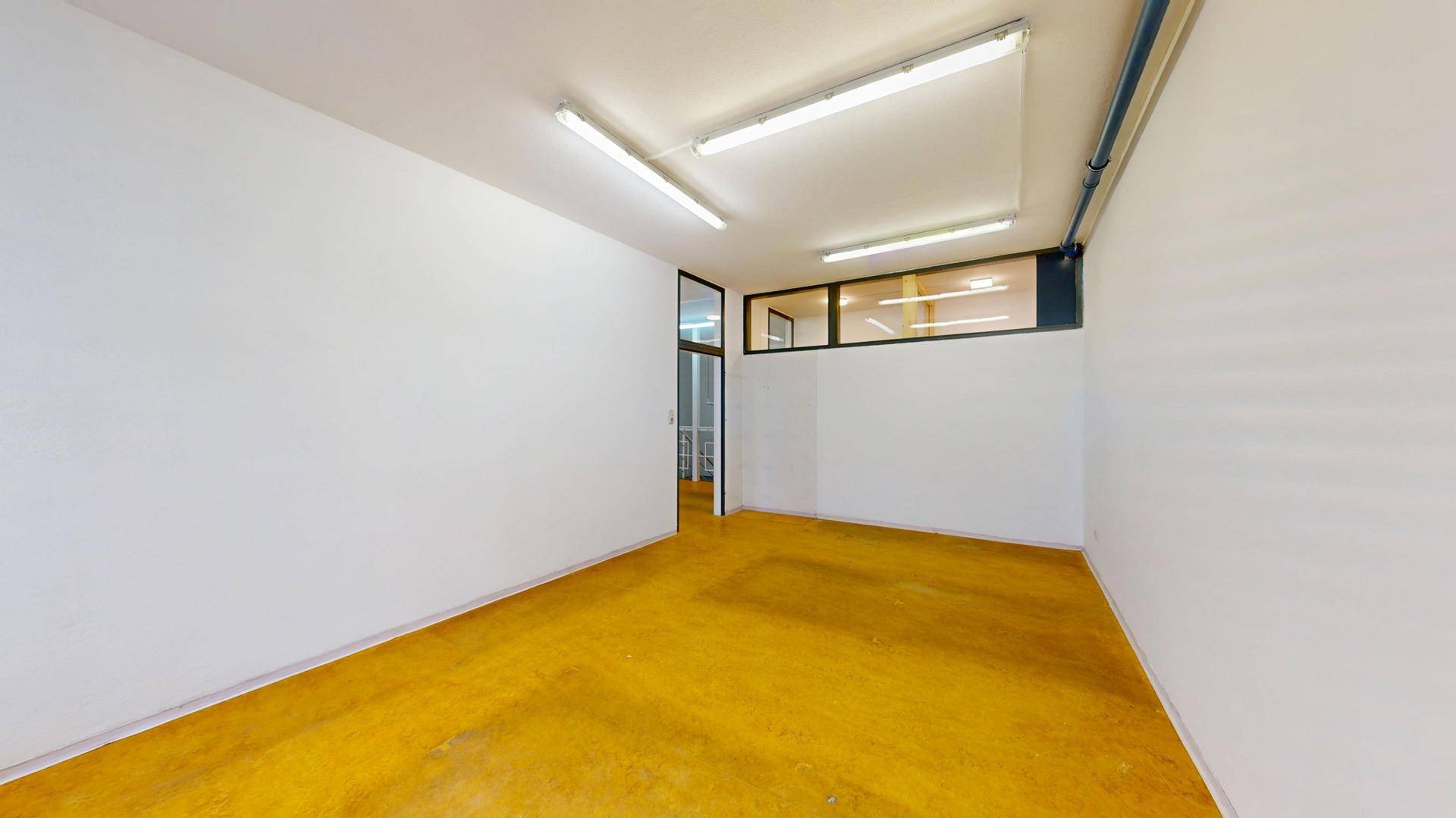 Heller Büroraum mit gelbem Boden, weißen Wänden und Zugang zu einem weiteren Bereich.