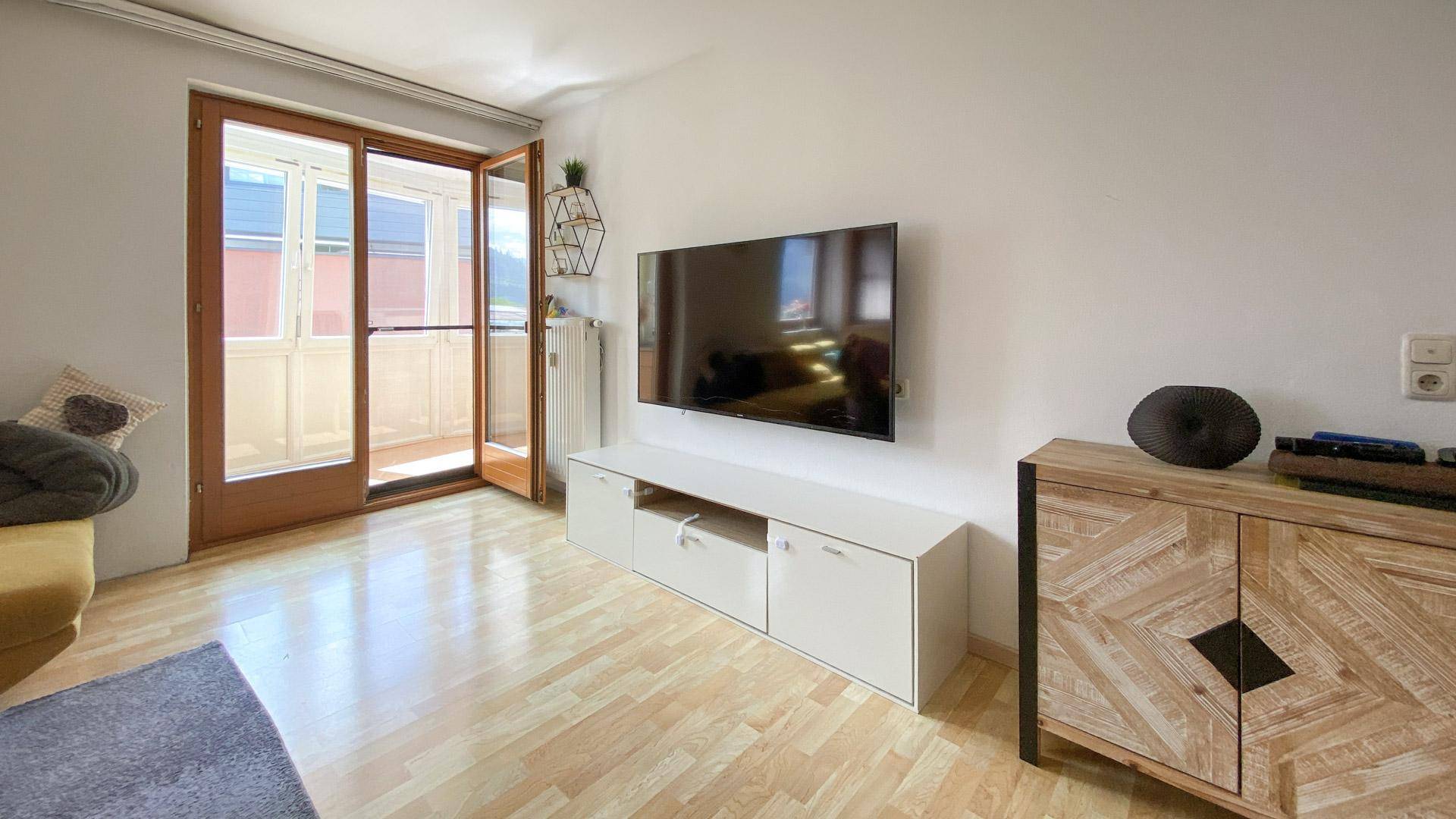 Wohnzimmer mit Wandfernseher, Sideboard und Zugang zu einem kleinen Balkon.