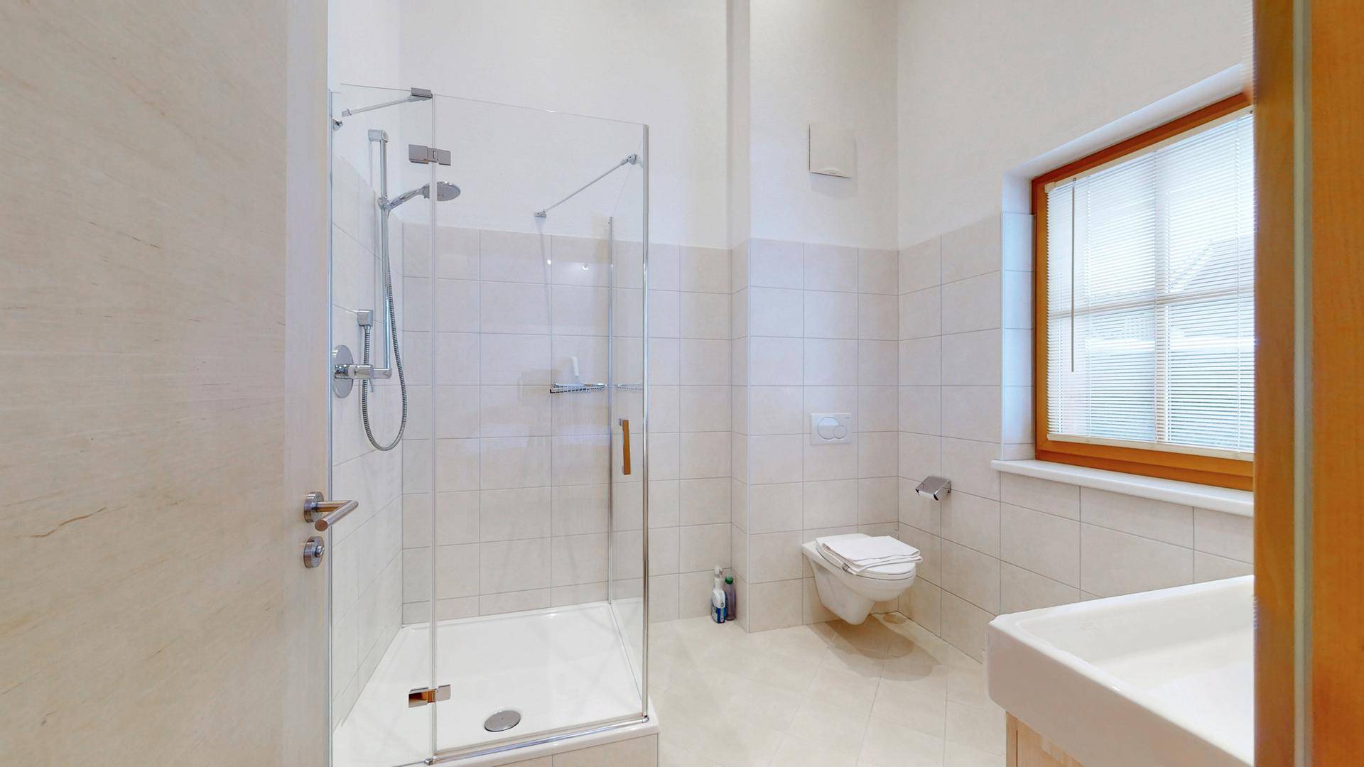 Helles Badezimmer mit Duschkabine, WC und Fenster mit Jalousien.