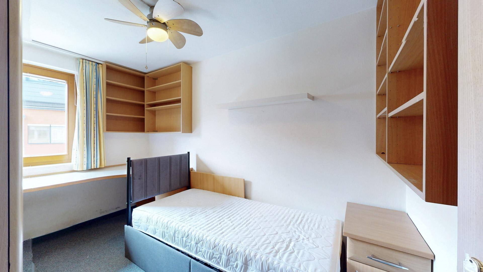 Schlafzimmer mit Einzelbett, integrierten Regalen und einem Schreibtisch am Fenster.
