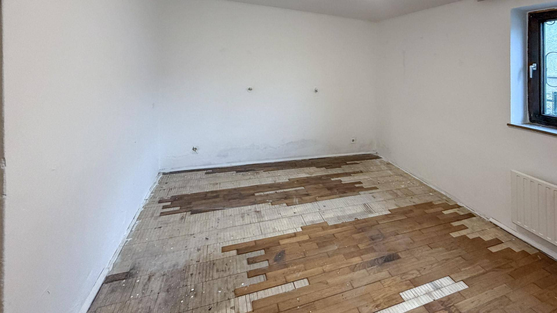 Leeres Zimmer mit beschädigtem Parkettboden und weißen Wänden, renovierungsbedürftig.