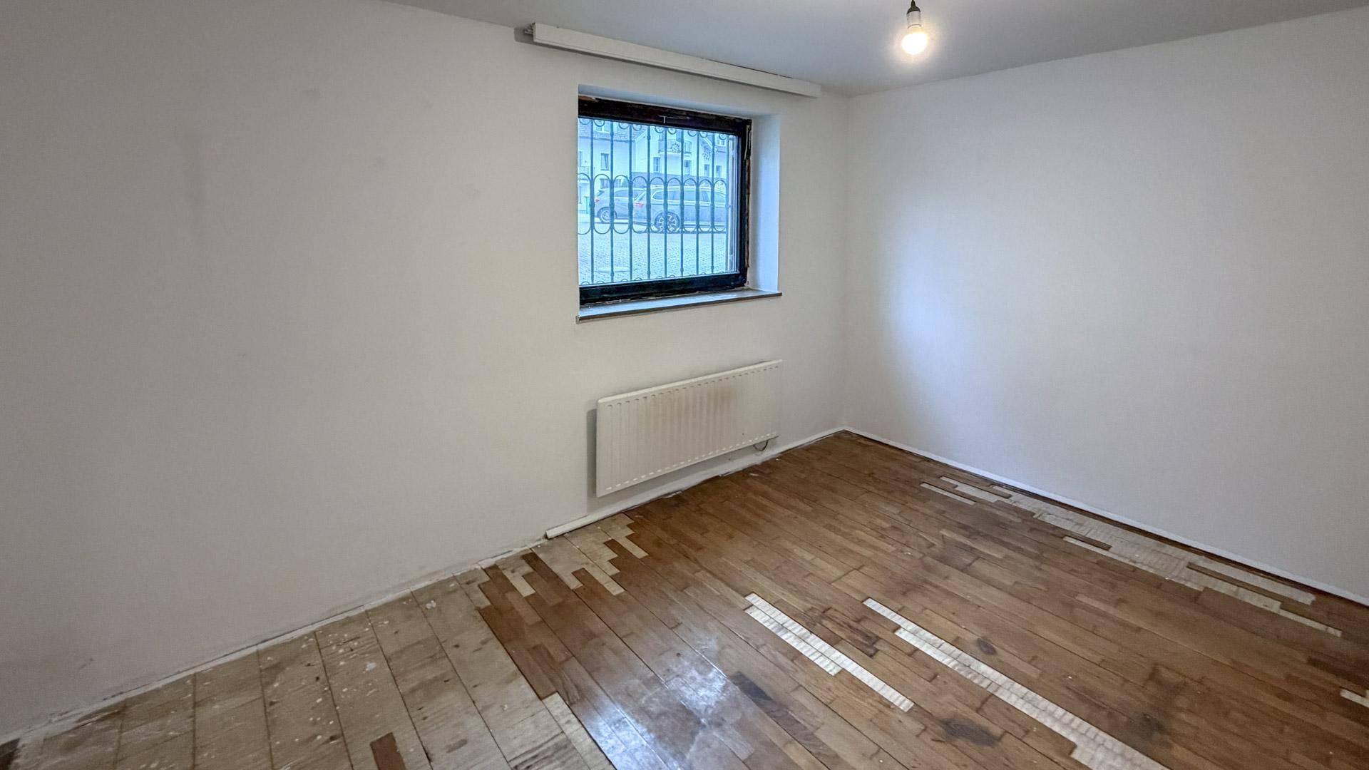 Leerer Raum mit Fenster, Heizkörper und beschädigtem Parkettboden, der Renovierung benötigt.
